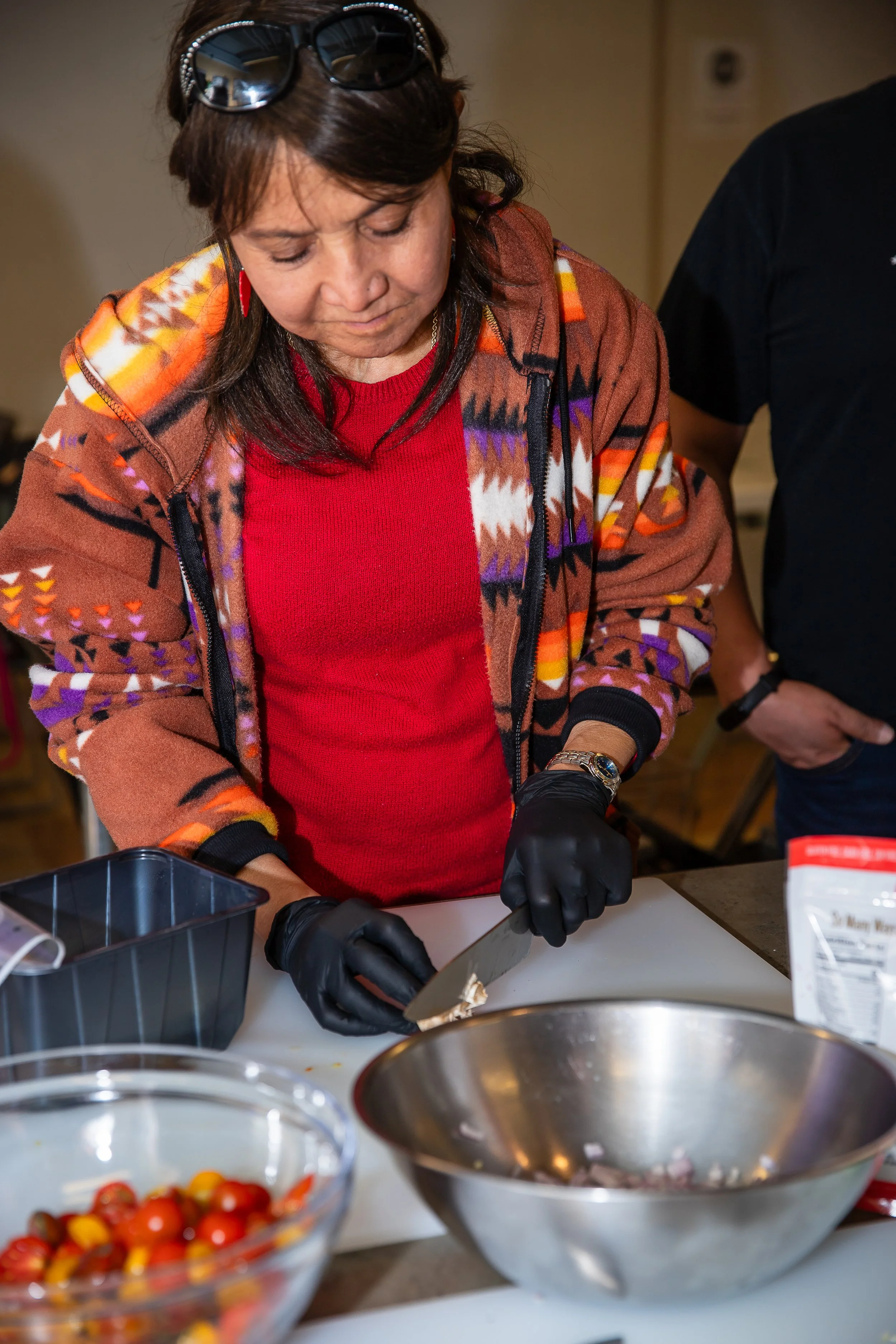 A woman wearing a colorful southwestern patterned jacket, red shirt, black gloves, and sunglasses on her head is chopping food on a white cutting board surrounded by bowls of colorful cherry tomatoes and other ingredients.