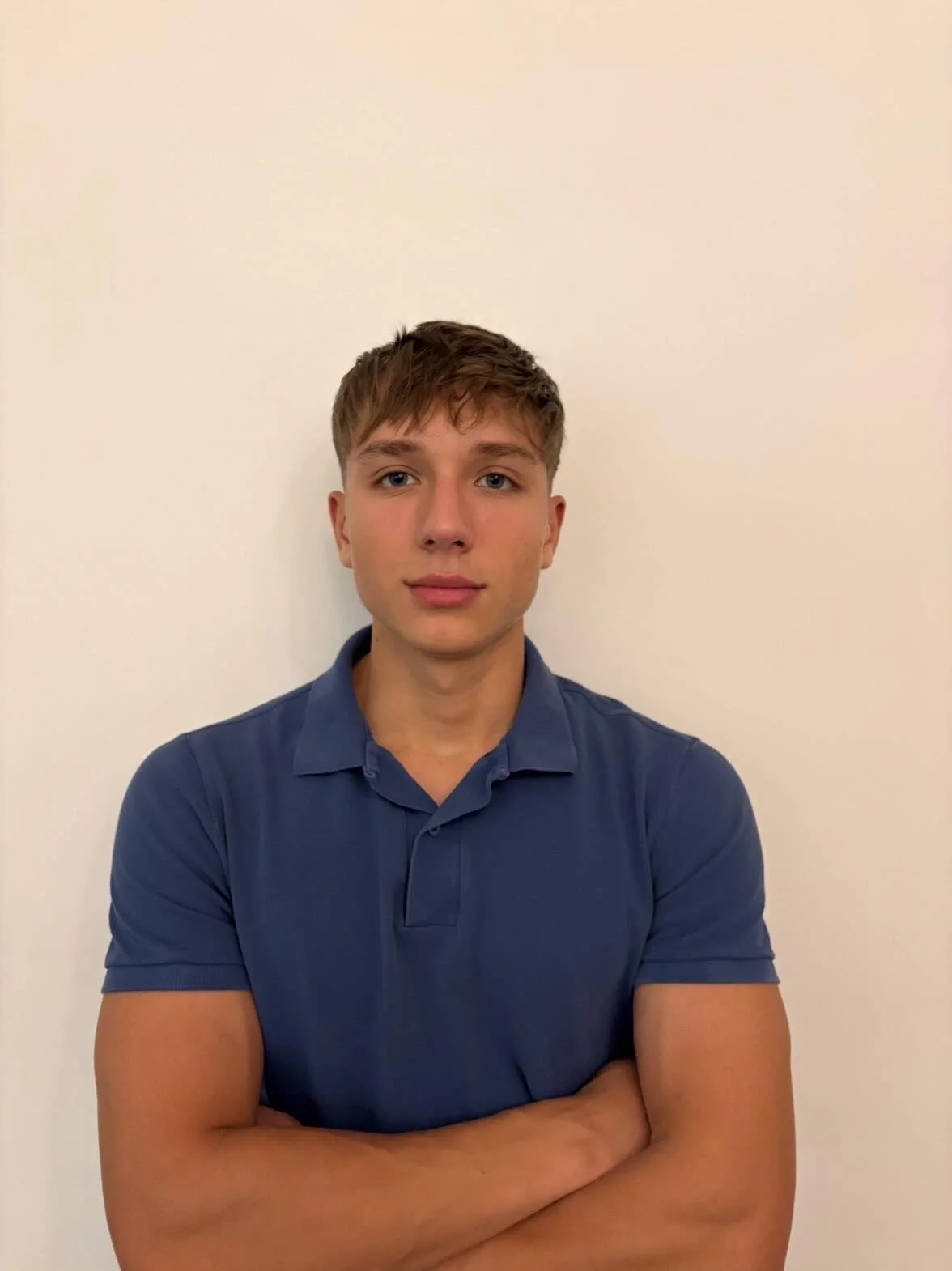 Young man with short light brown hair and blue eyes, wearing a blue polo shirt, standing against a plain white wall with arms crossed.