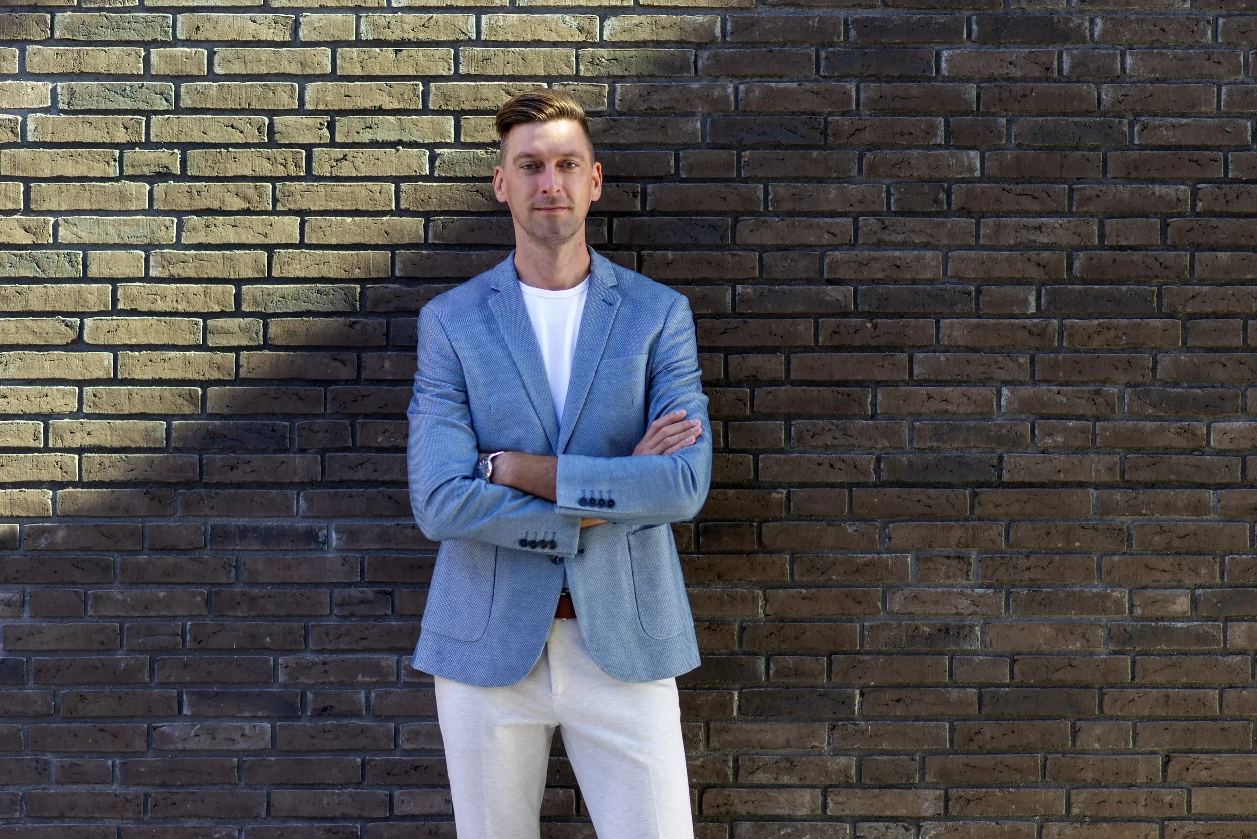 A man standing with arms crossed in front of a brick wall, wearing a light blue blazer, white shirt, and beige pants.