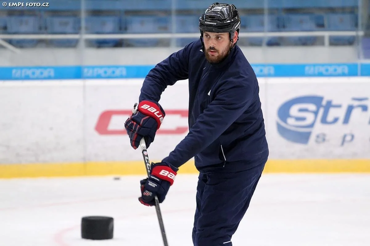 A man with a beard in hockey gear practicing on an ice rink, holding a hockey stick, with a puck nearby.