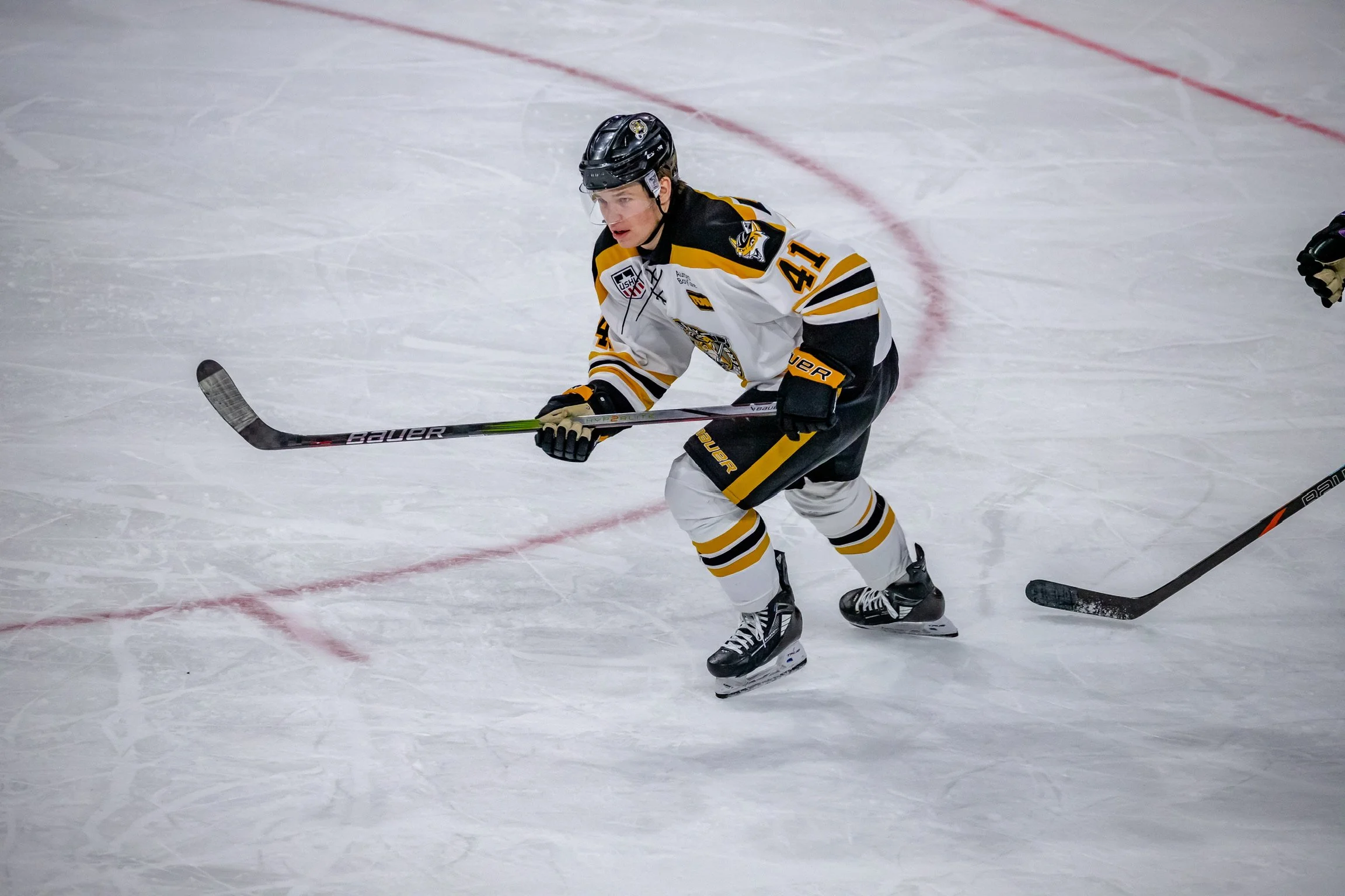 A hockey player in a black, white, and yellow uniform is on the ice, handling the puck with a hockey stick, wearing a helmet and skates.