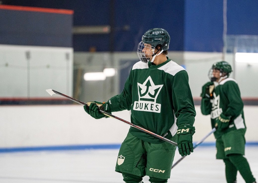 Two ice hockey players in green jerseys practicing on the ice rink, with one player in the foreground holding a hockey stick and the other in the background.