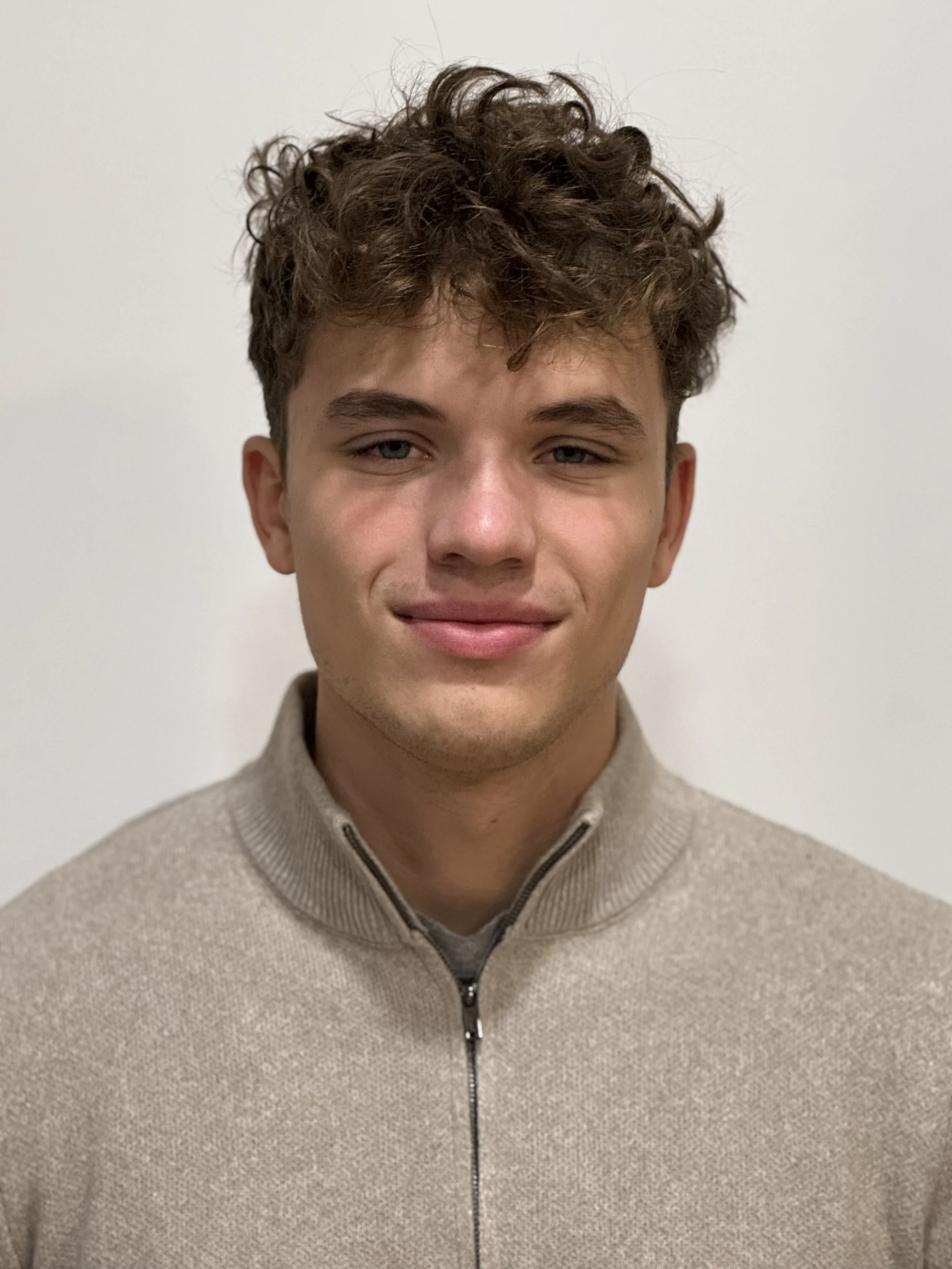 A young man with curly brown hair and fair skin, smiling slightly, wearing a beige zip-up sweater against a plain white background.
