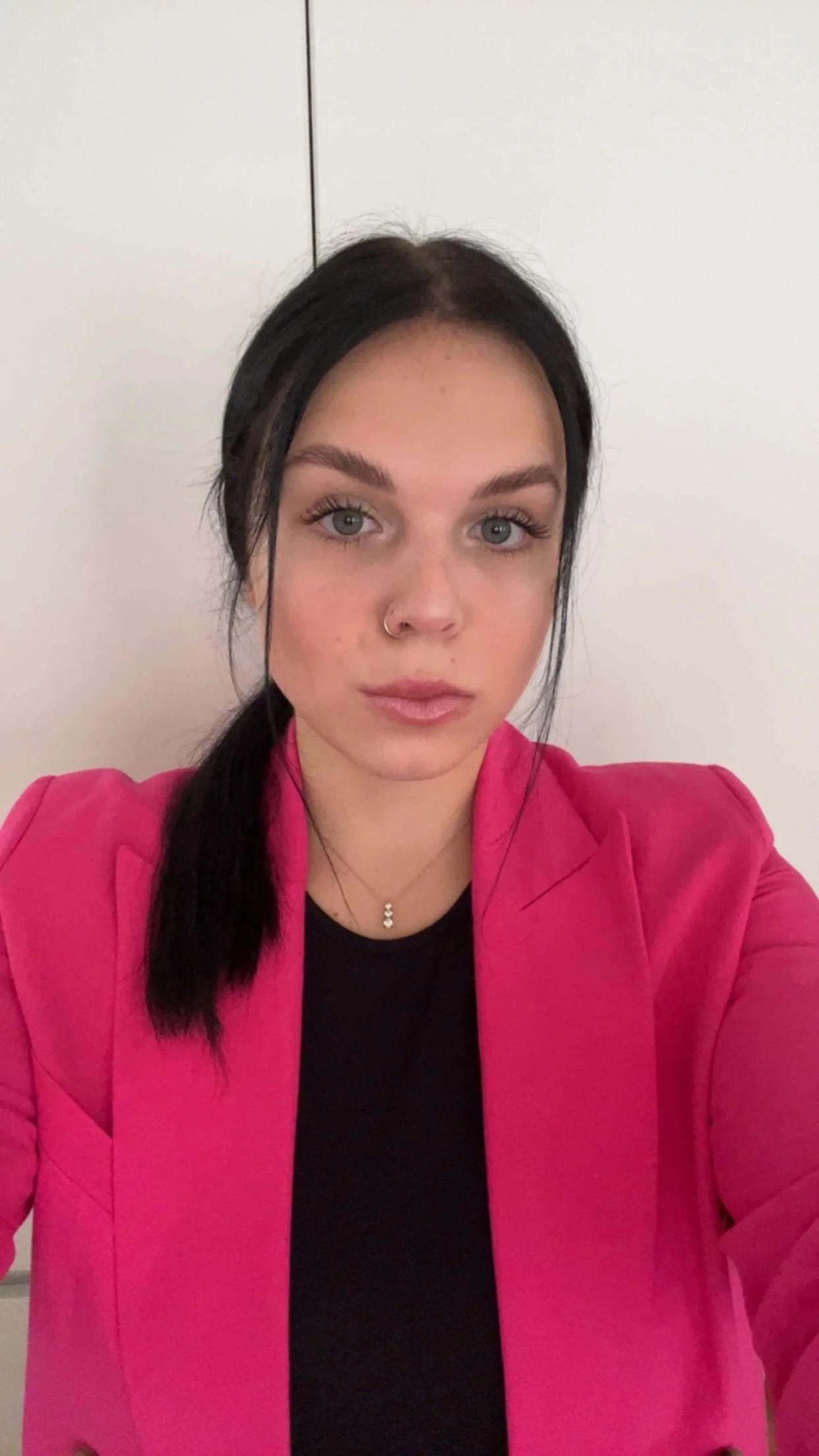 A young woman with blue eyes and dark hair in a ponytail, wearing a pink blazer over a black top, standing against a plain white background.