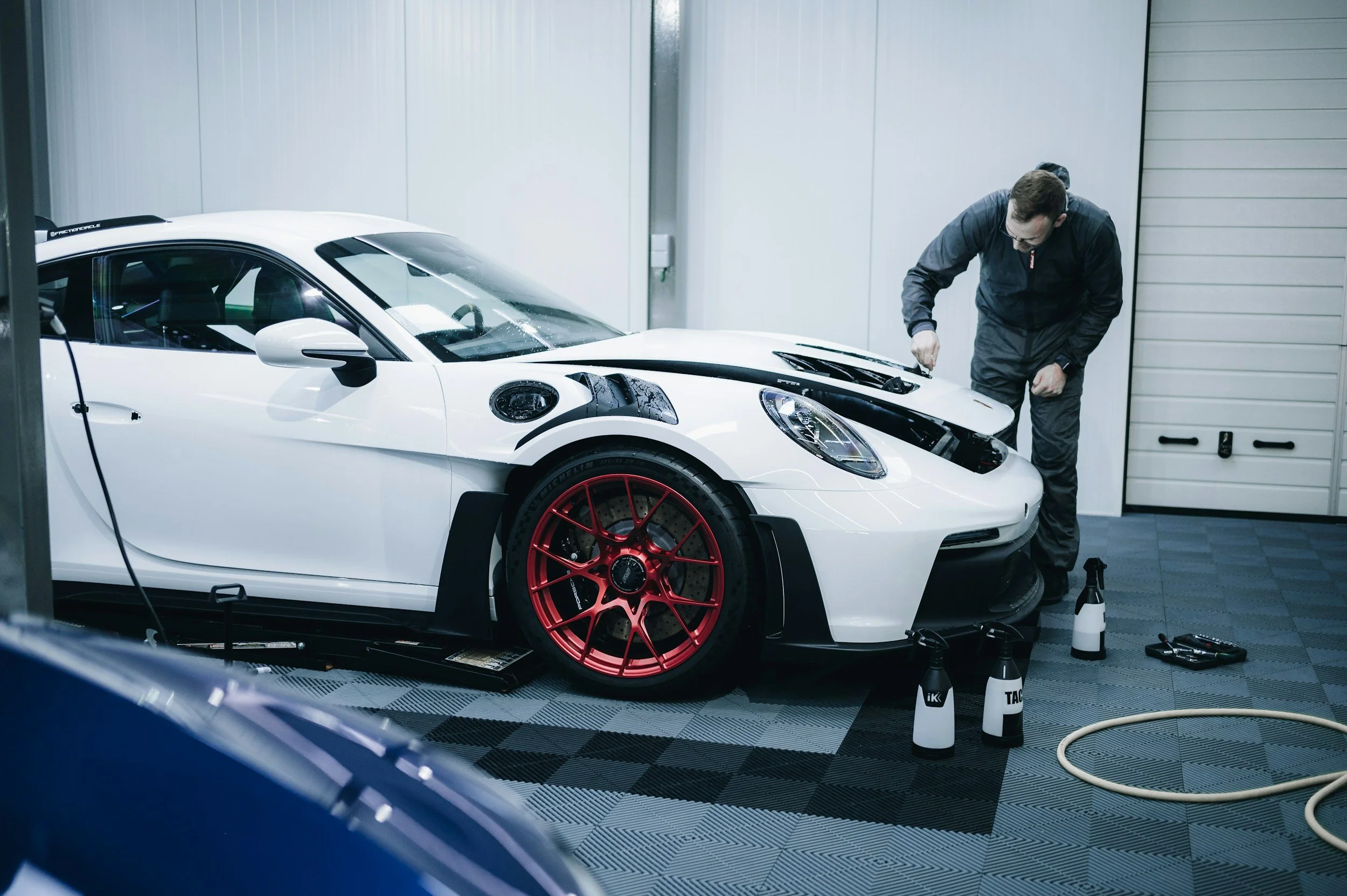 A mechanic working on a white sports car with red wheels in a garage, with tools and spray bottles nearby.