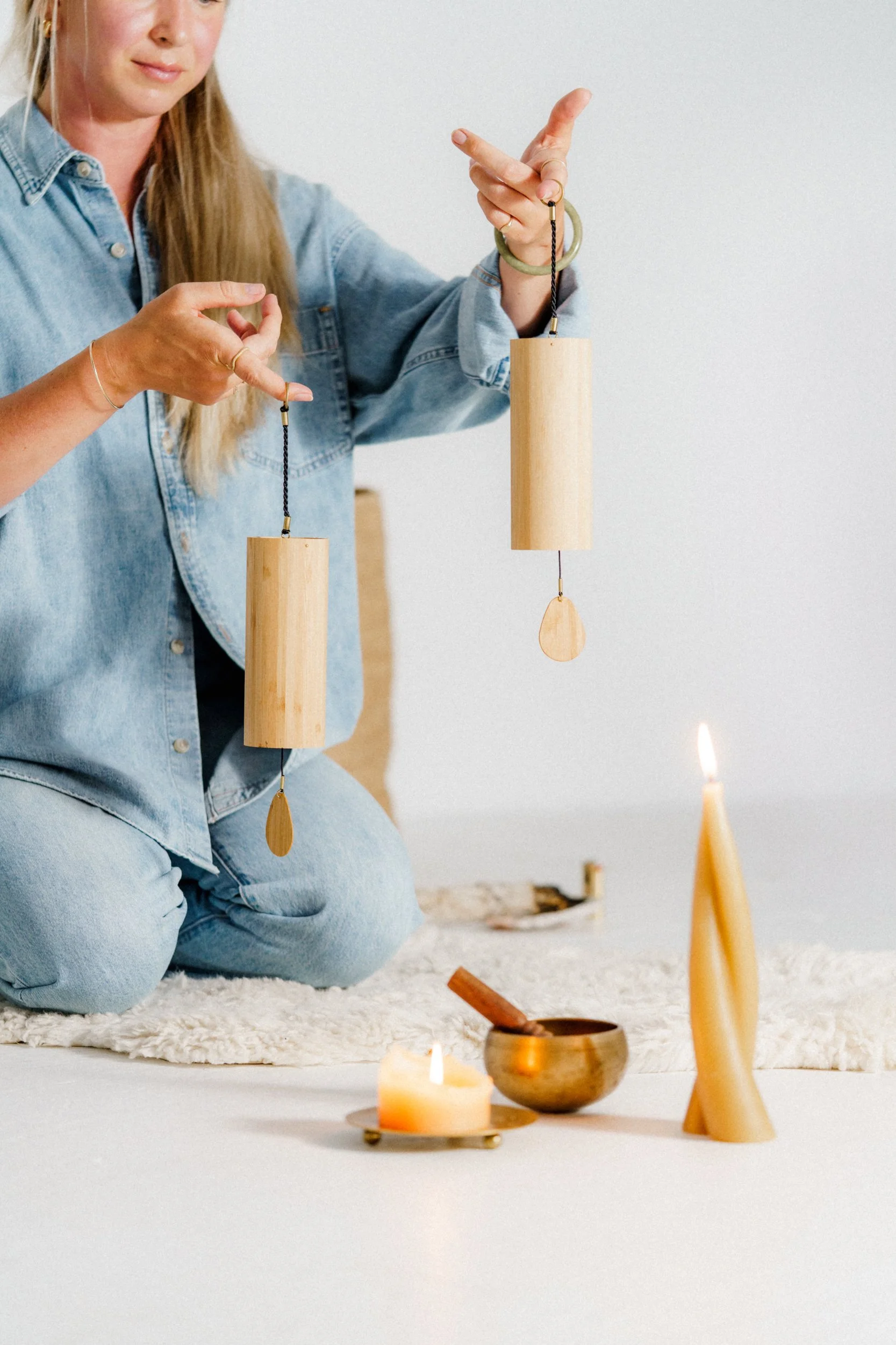 A woman in a denim shirt and jeans kneeling on a white rug, holding two wooden wind chimes with lit candles and a singing bowl on the floor in front of her.