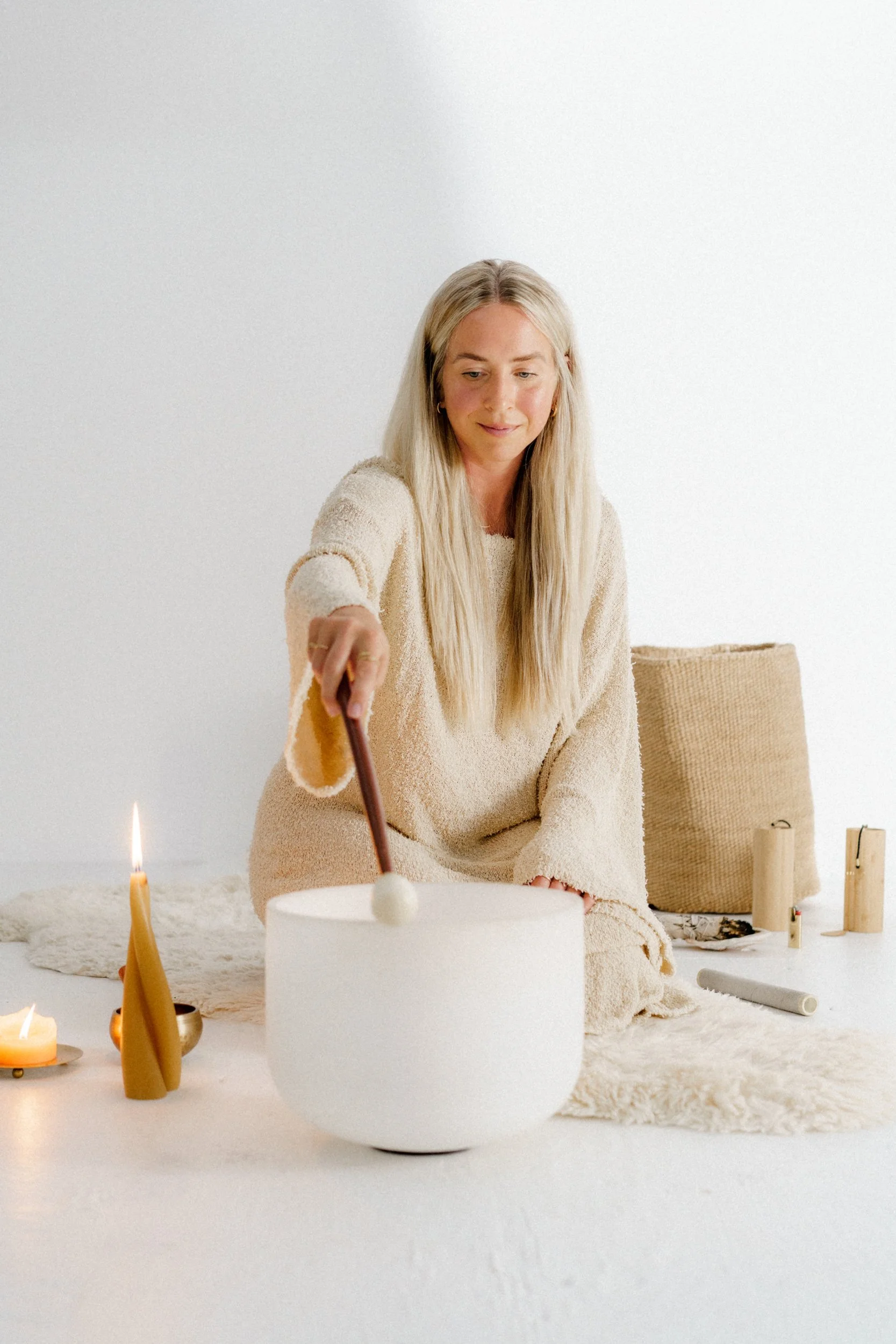 A woman with long blonde hair in a beige sweater playing a crystal singing bowl surrounded by candles and wooden instruments in a minimalist, cozy space.