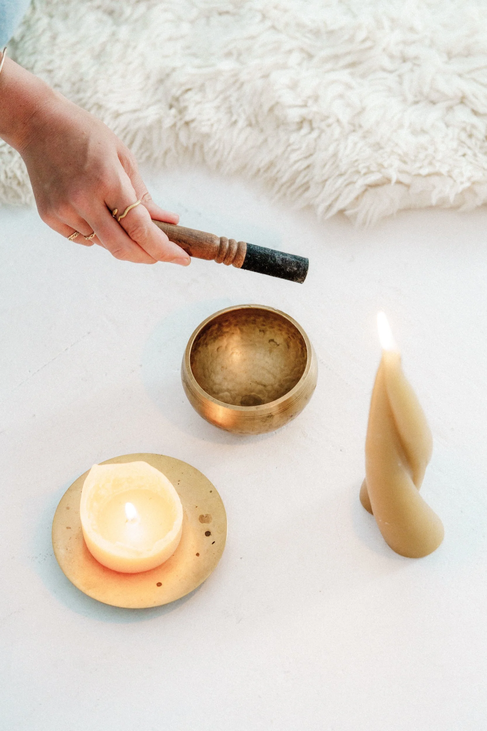 Person ringing a singing bowl and two partially melted candles on a white surface with a white furry blanket in the background.