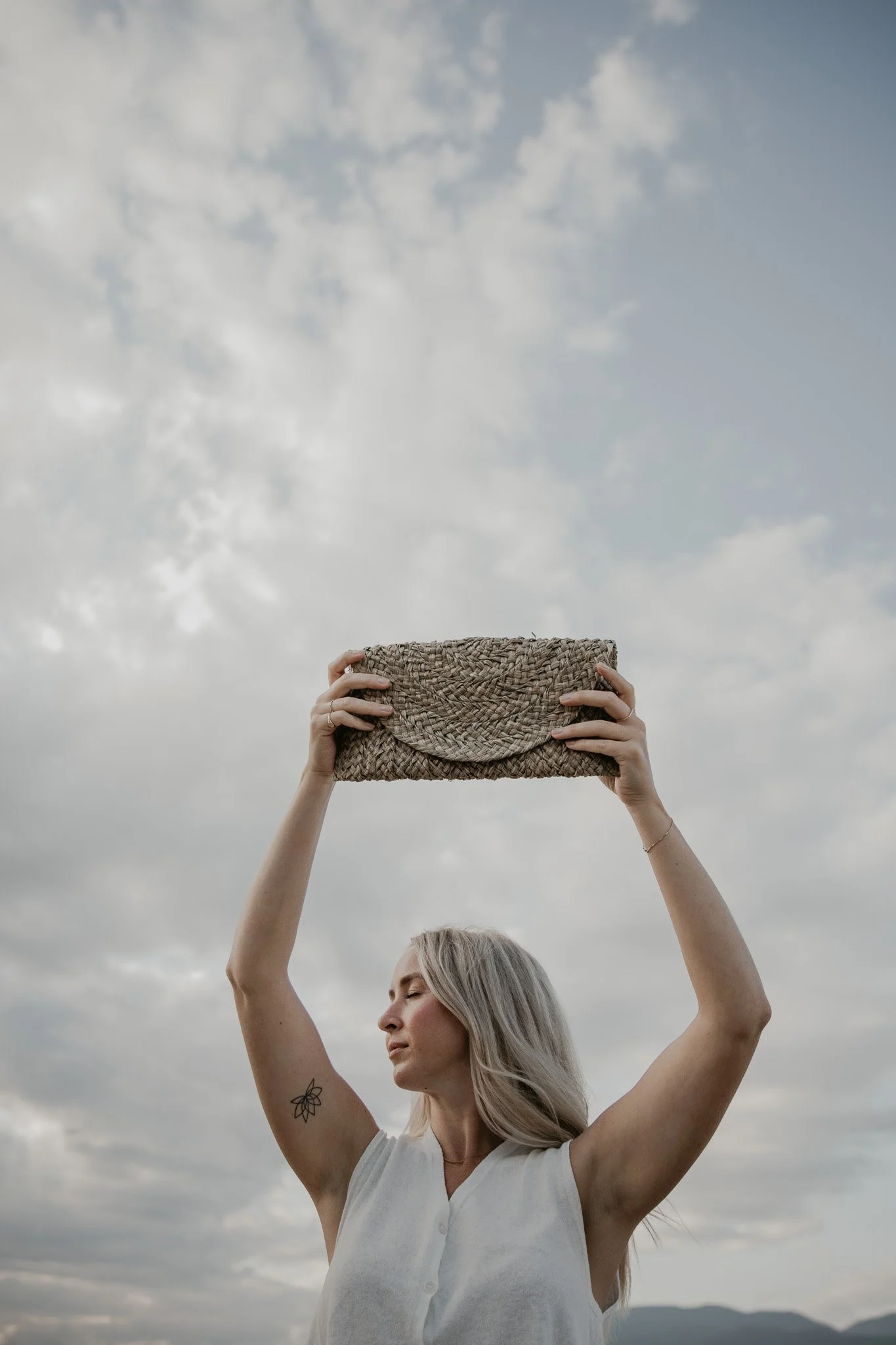 A woman with blonde hair, wearing a sleeveless white top, holds a woven clutch above her head against cloudy sky backdrop.