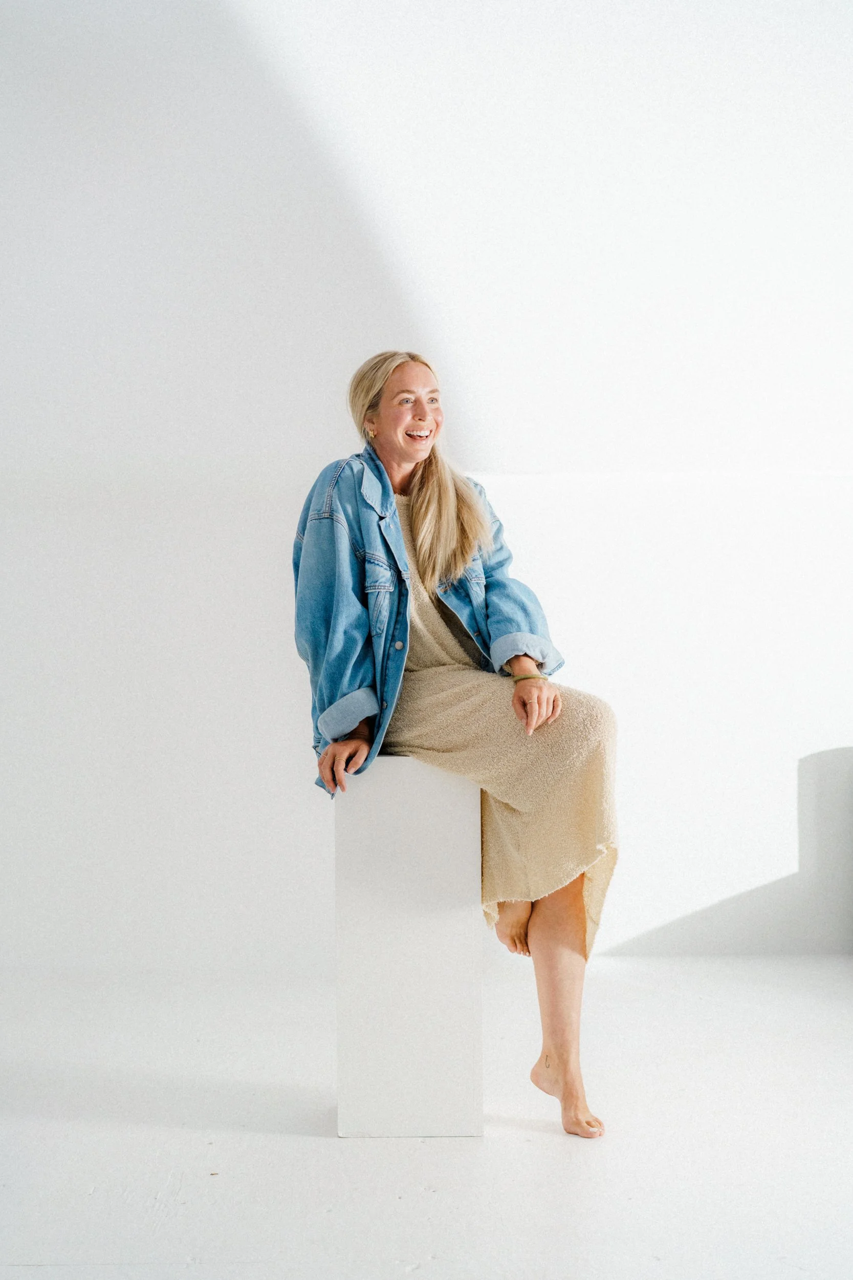 A woman with long blonde hair wearing a beige dress and denim jacket, sitting barefoot on a white cube in a minimalist white studio, smiling and looking to the side.