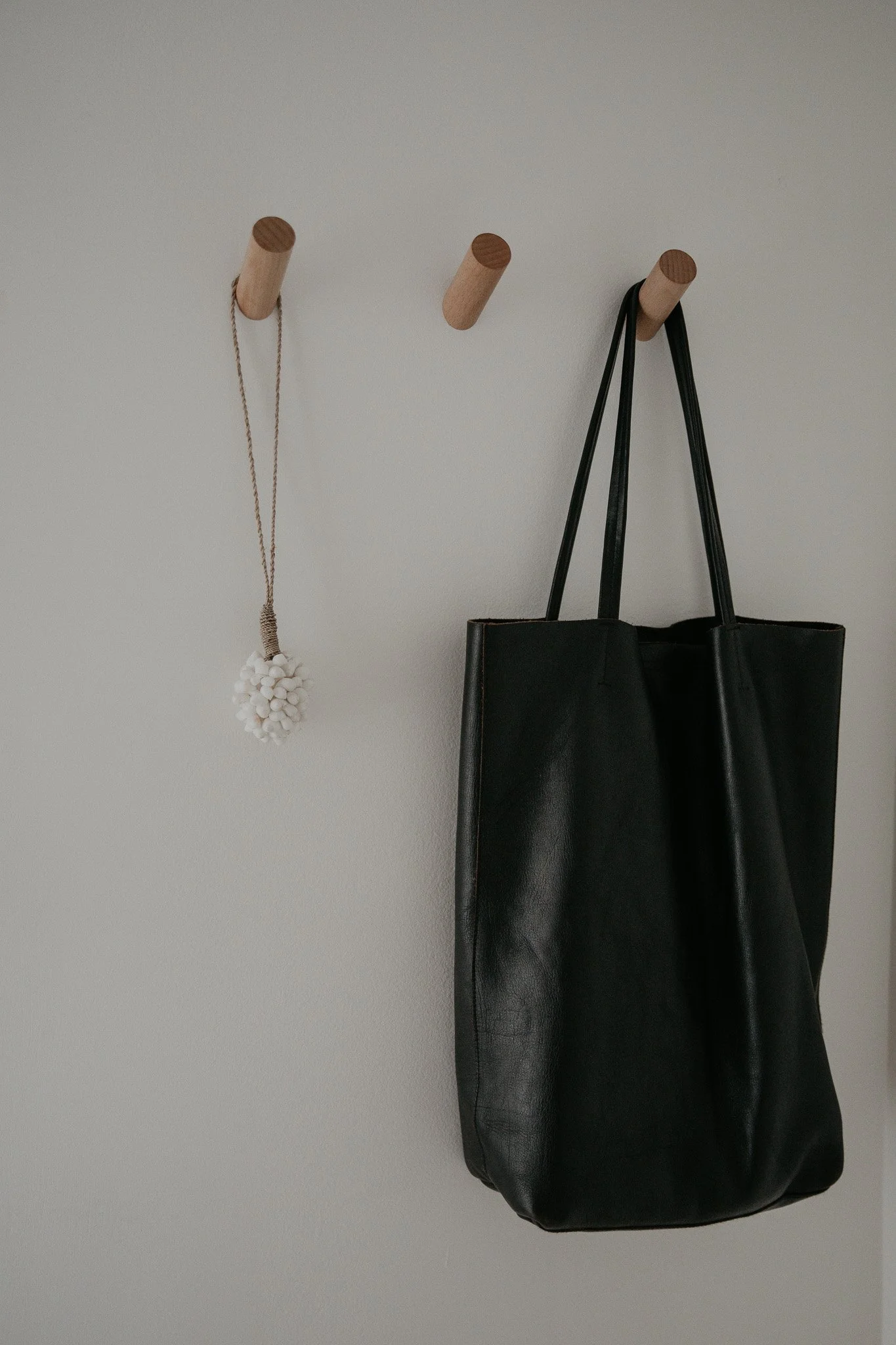 A wall with three wooden hooks, one holding a white beaded necklace, and another holding a black leather tote bag.