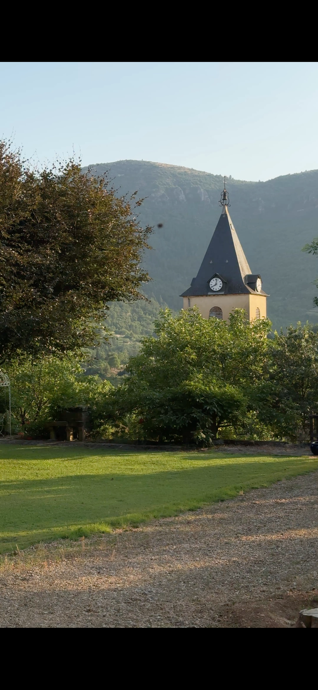 A church with a clock on its steeple is visible through green trees, mountains in the background, and a grassy area in the foreground.
