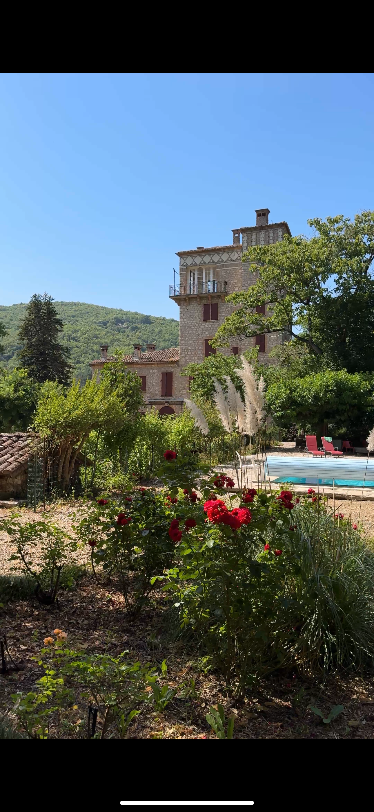 A scenic view of a stone house surrounded by lush greenery, flowering plants, a swimming pool, and a mountain in the background on a sunny day.