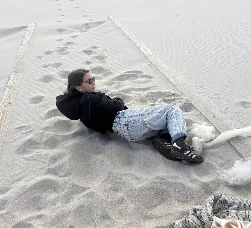 Person lying on a sandy surface in a small, sealed area surrounded by wooden borders, with visible footprints and a plastic bag nearby.