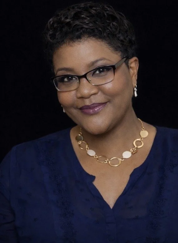 Portrait of a woman with short curly black hair, glasses, and wearing a blue top, with gold and white jewelry, against a black background.