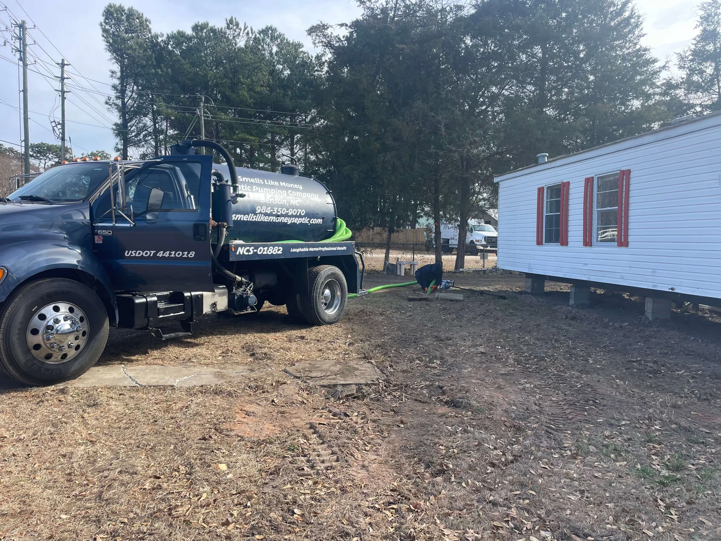 Septic truck parked near a white mobile home with red shutters, with a worker inspecting the septic tank access in a yard with sparse grass and dirt.