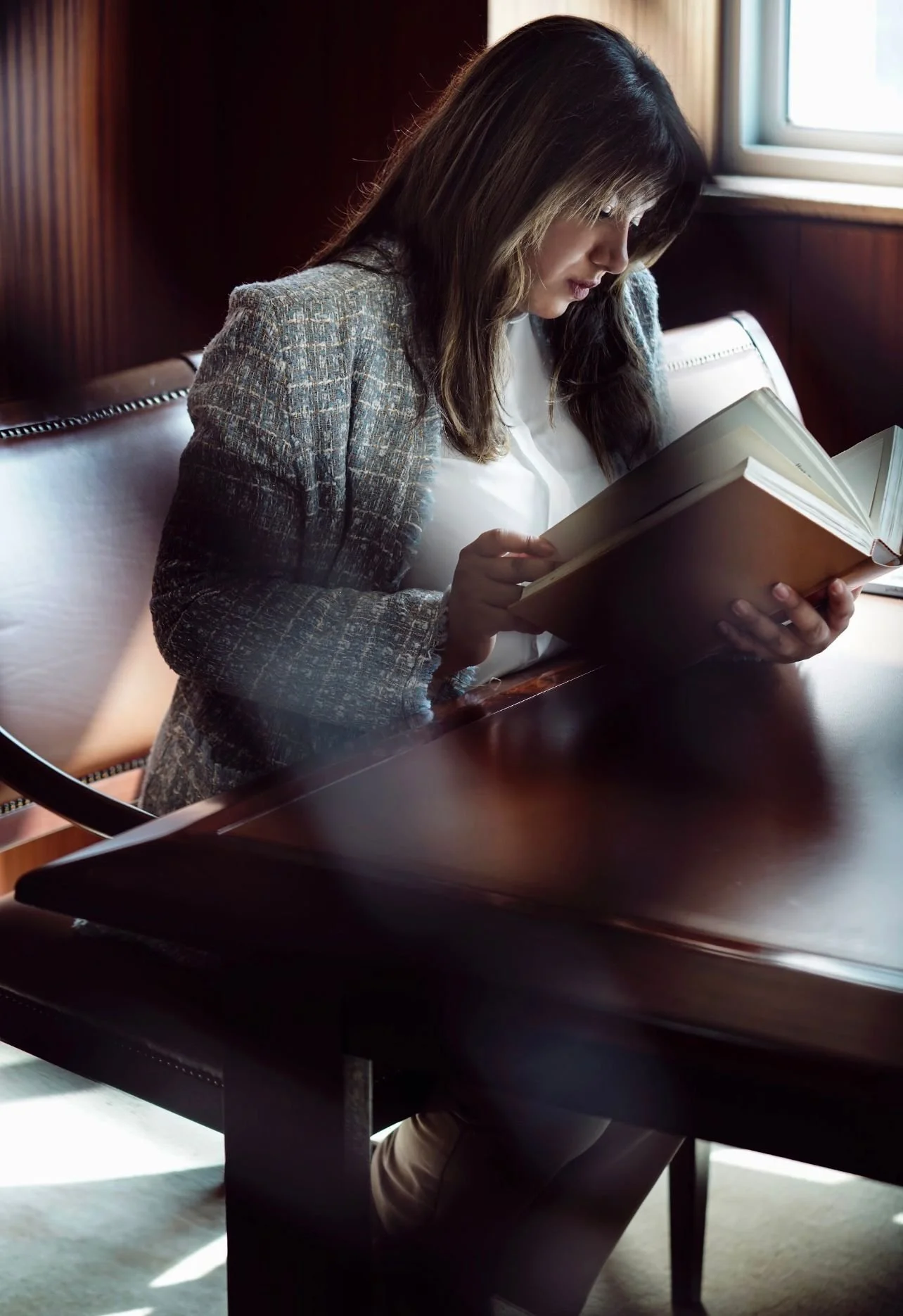 A woman with long brown hair reading a book while sitting at a dark wooden table near a window.