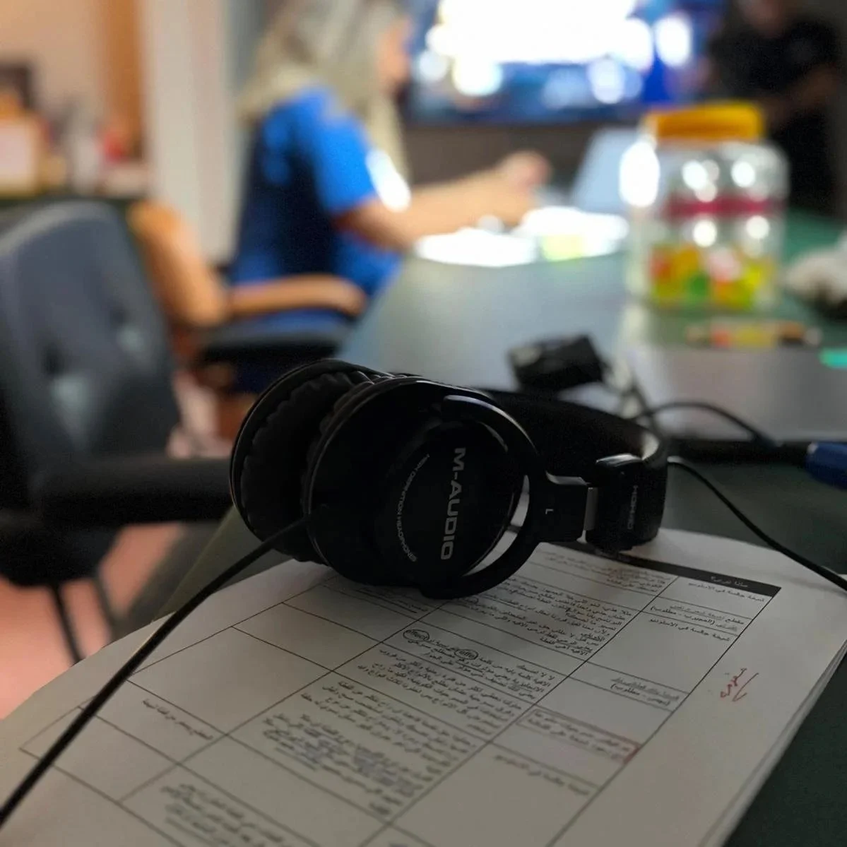 Person holding a handwritten paper with Arabic script, standing inside a room with a round table, rug, and camera equipment.