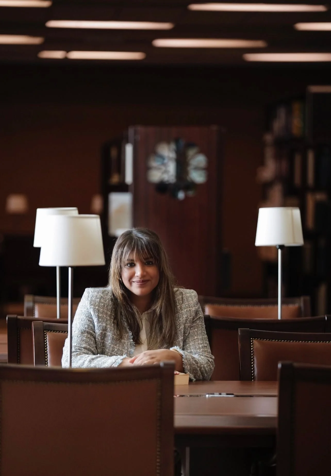 A woman with long brown hair and a beige blazer sitting at a wooden table in a dimly lit room, with two white lamps and a bookshelf in the background.