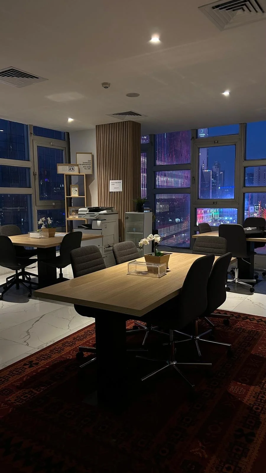An office conference room with a wooden table, black chairs, a flower arrangement, and city lights visible through the large windows at night.