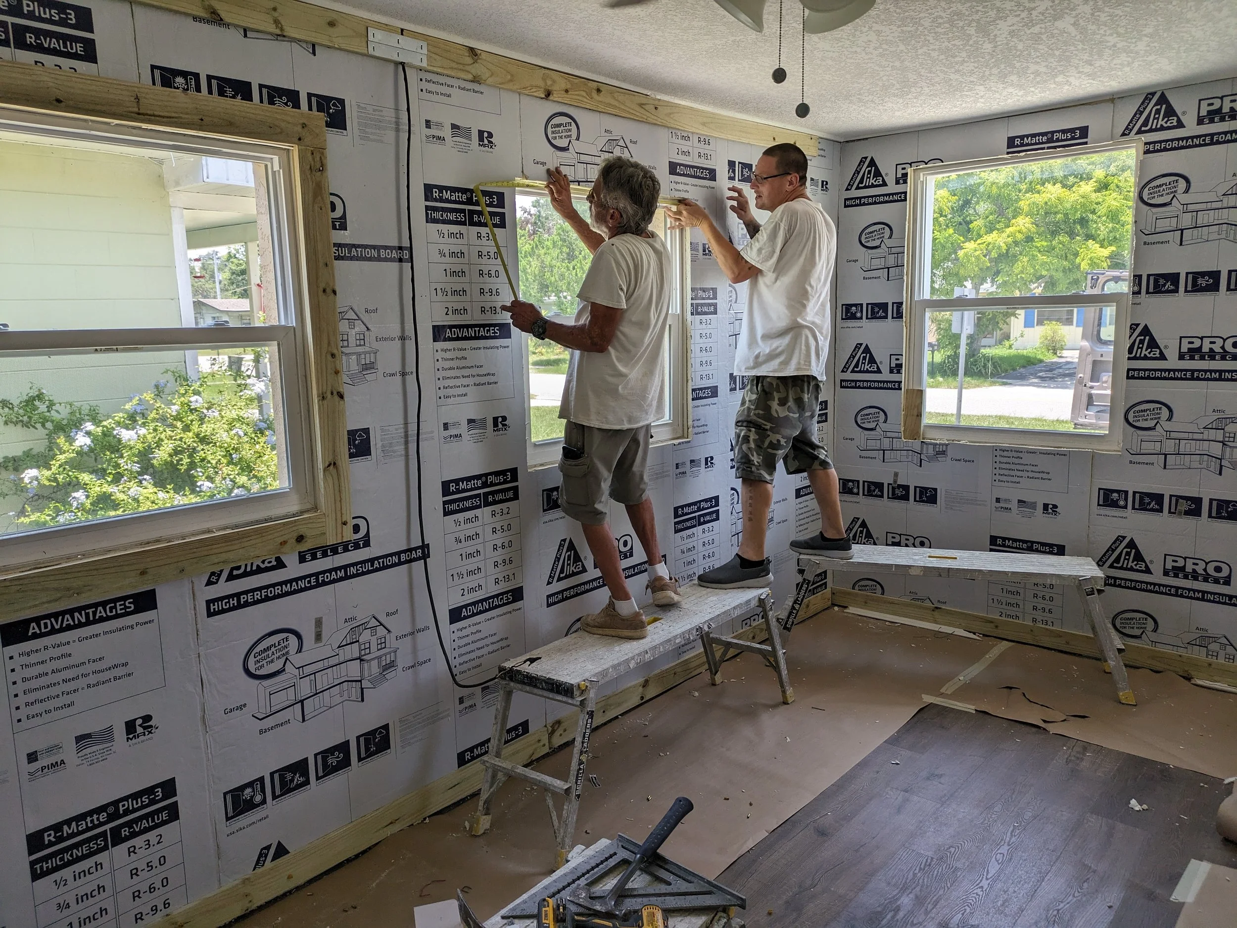 Two men working on interior insulation installation in a room with windows, standing on ladders and using tools to attach insulation boards to the walls.