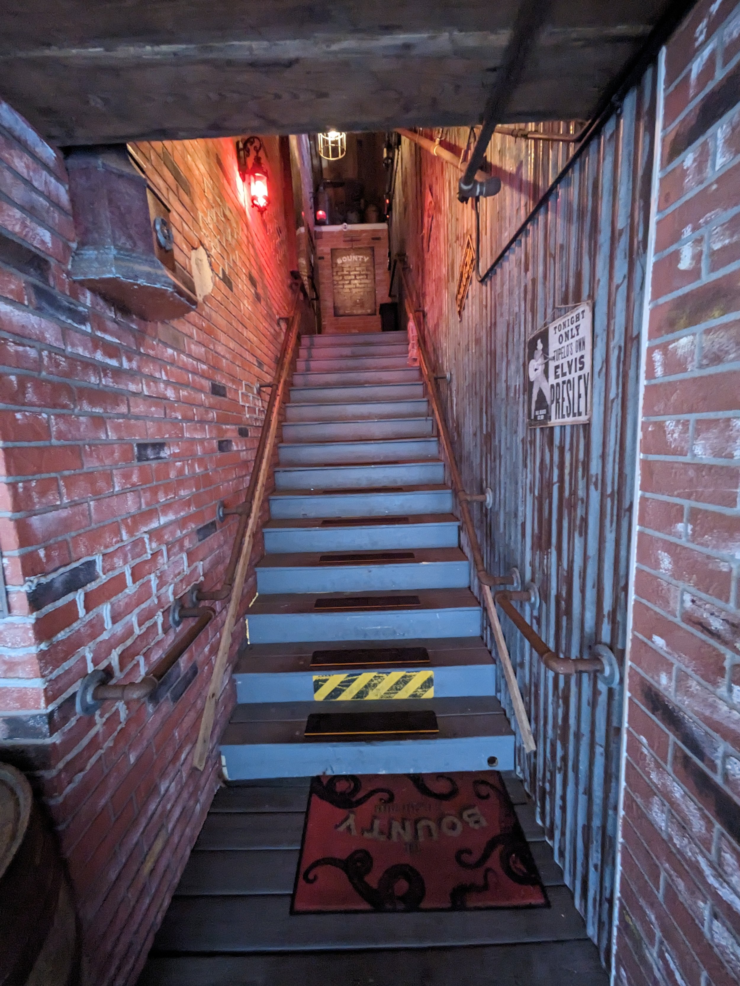 A narrow staircase with metal steps and wooden handrails, surrounded by red brick walls and corrugated metal panels, leading upward in a dimly lit bar or pub setting with vintage signs, lights, and a Welcome mat at the bottom.