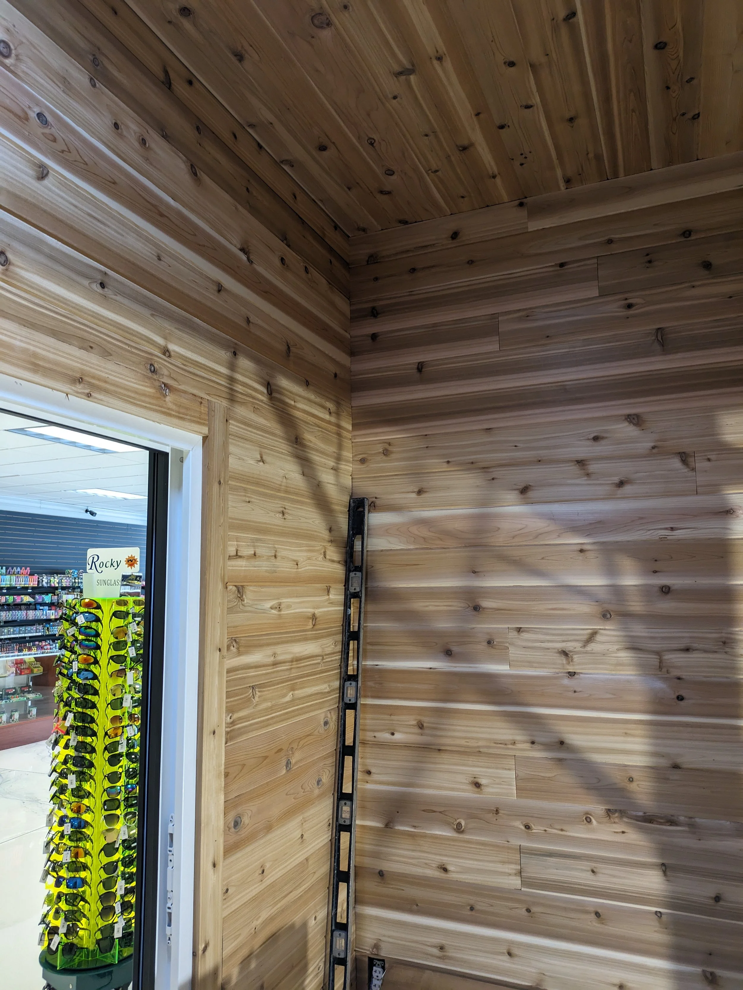 Empty corner of a wooden-paneled room with a leveling tool and an outlet on the wall, next to a door leading to a store with sunglasses display.