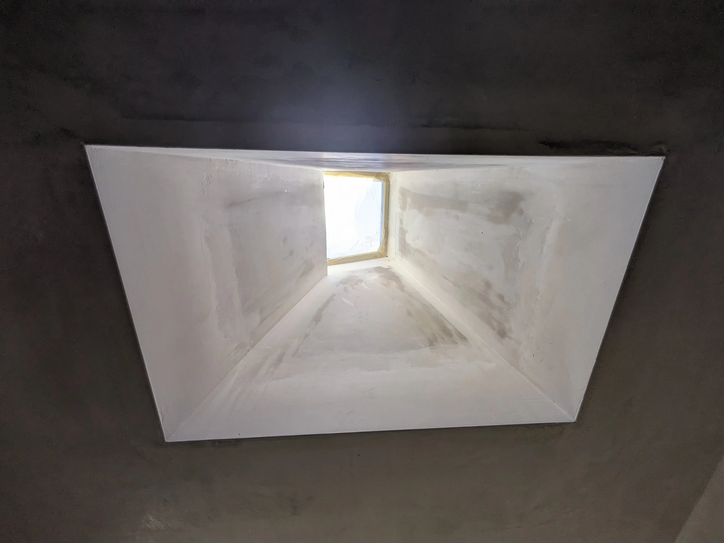 Looking up through a rectangular attic or ventilation hatch with a clear window at the top, illuminated by natural light.