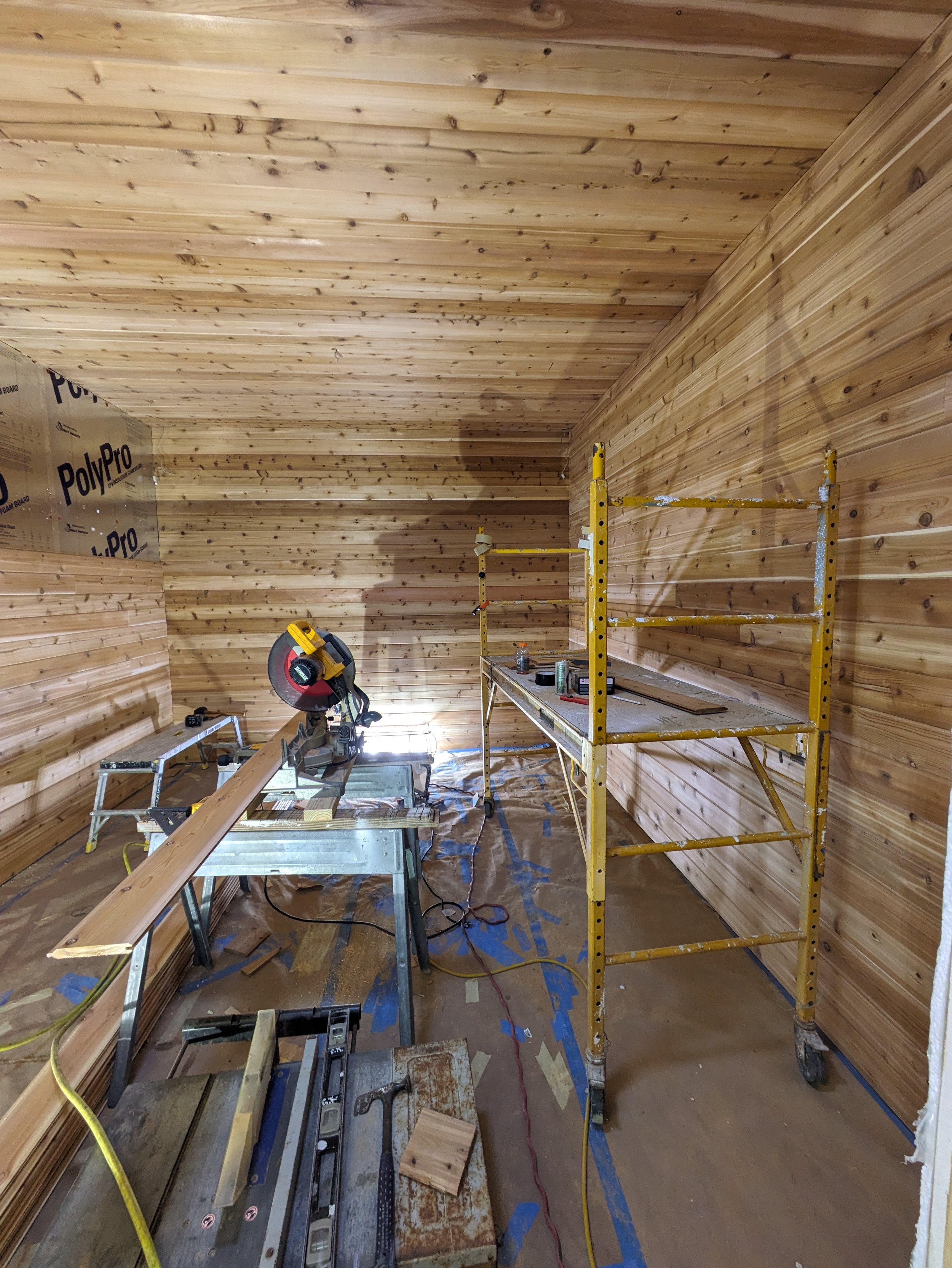 Room under construction with wooden walls and ceiling, yellow scaffolding, saw on table, and construction tools scattered on the floor.