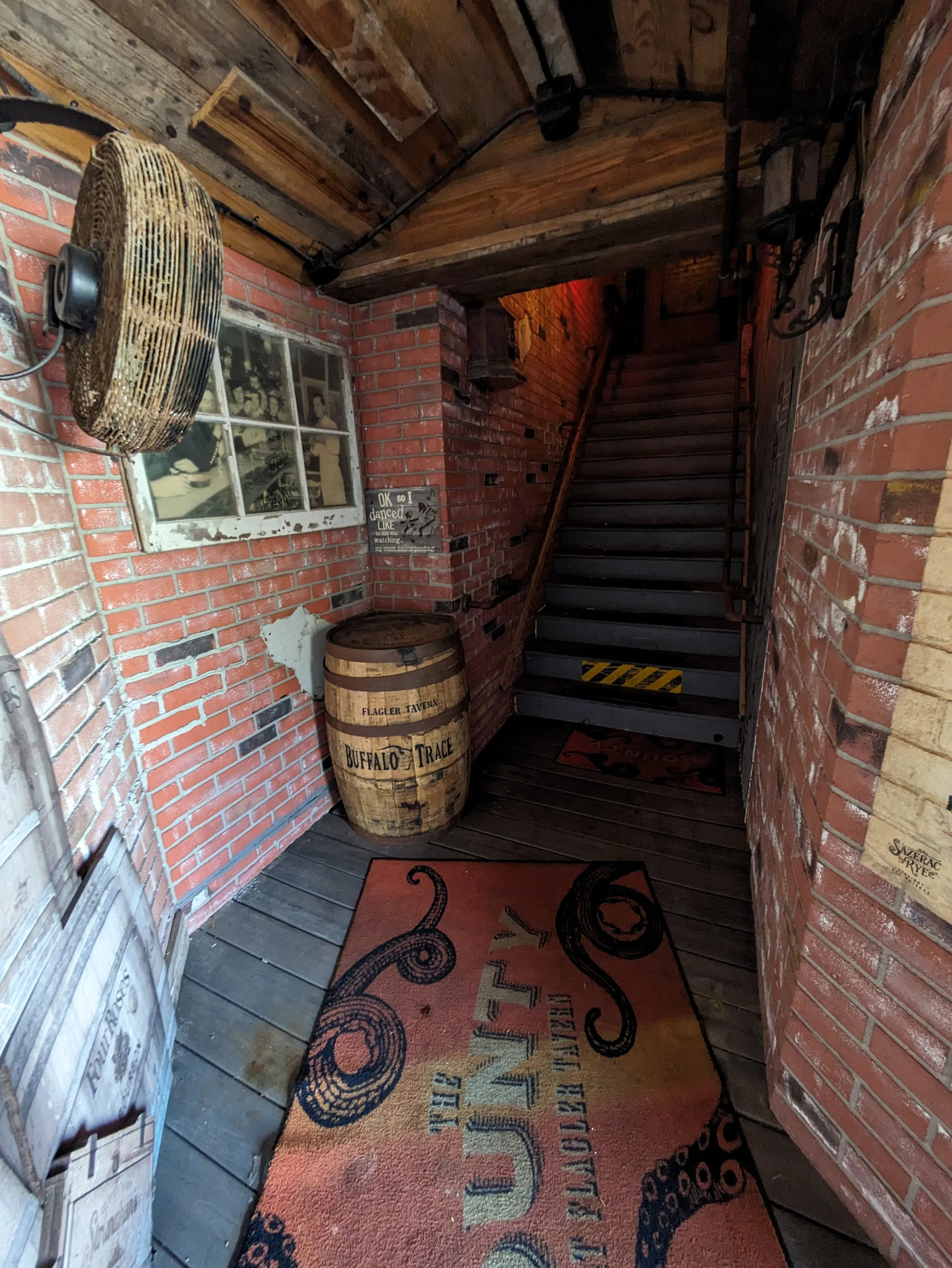 Interior of a rustic bar or tavern with exposed brick walls, a wooden staircase, a barrel labeled Buffallo Trace, a wall fan, and a colorful welcome mat.
