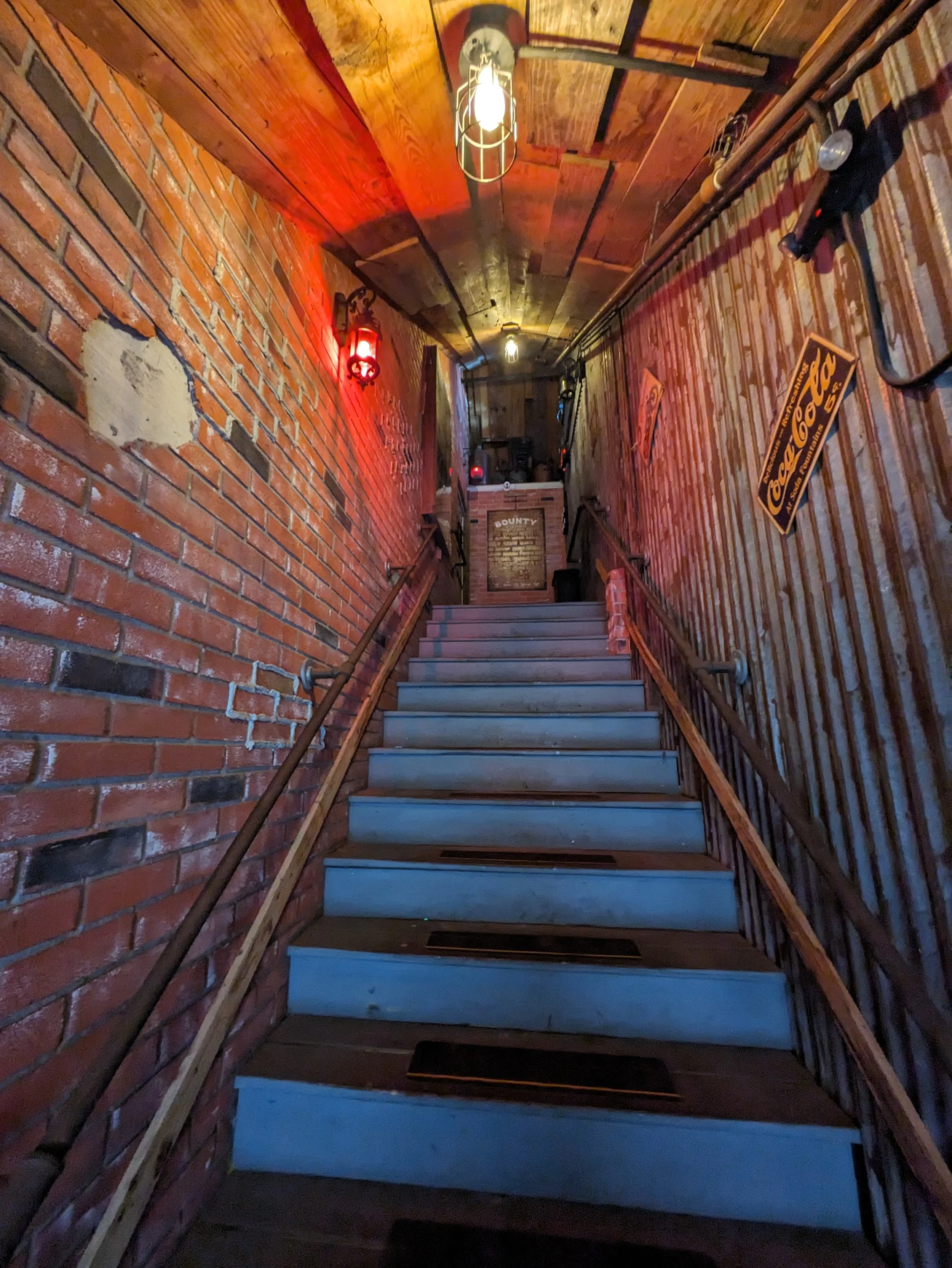 A dimly lit stairway with wooden steps and metal railings, leading to a brick and wood interior with vintage signs and exposed lighting fixtures.