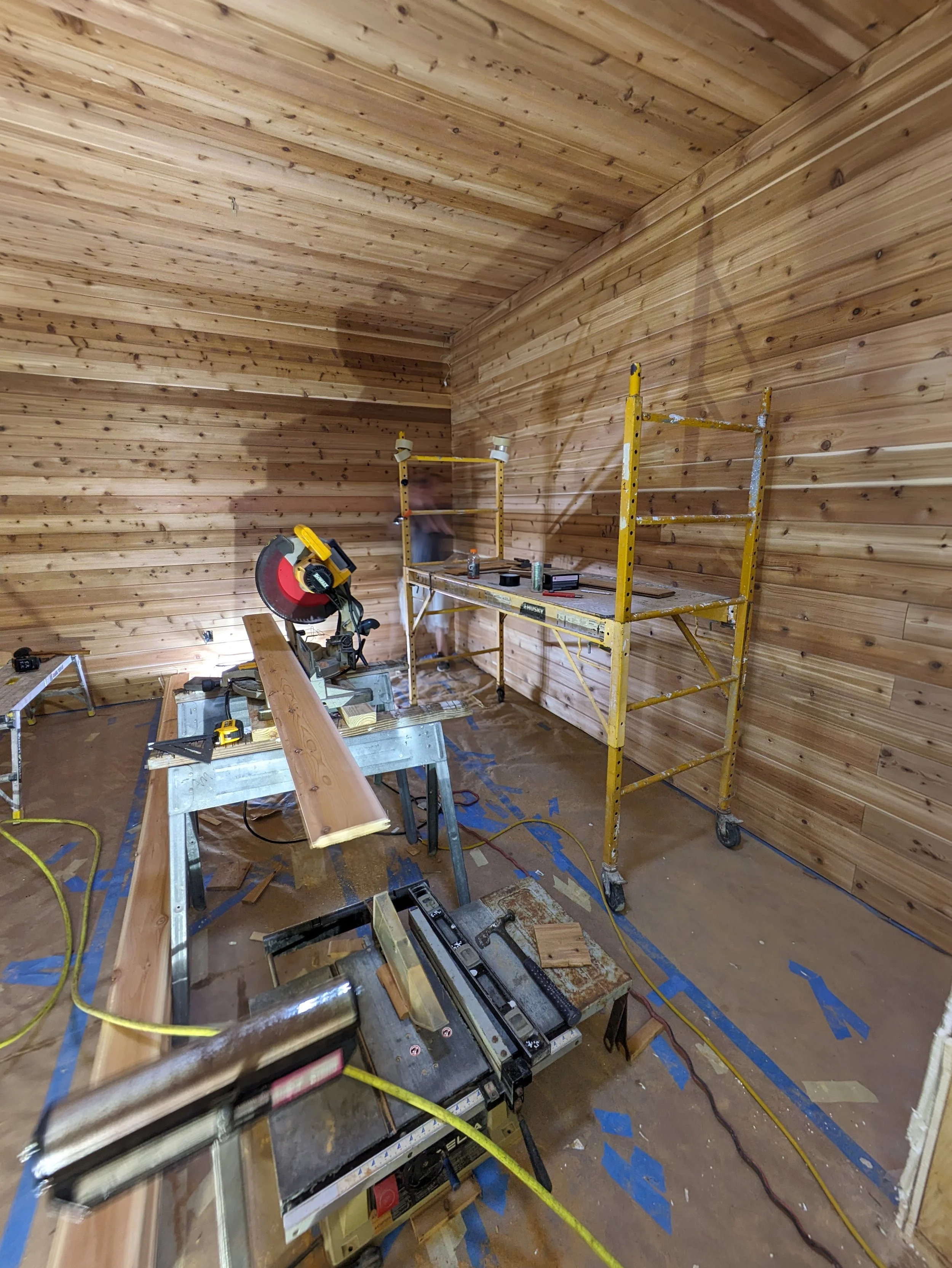 Interior of a woodworking workshop with wood-paneled walls, a miter saw on a workbench, a yellow scaffold, and various tools and wood pieces scattered around.