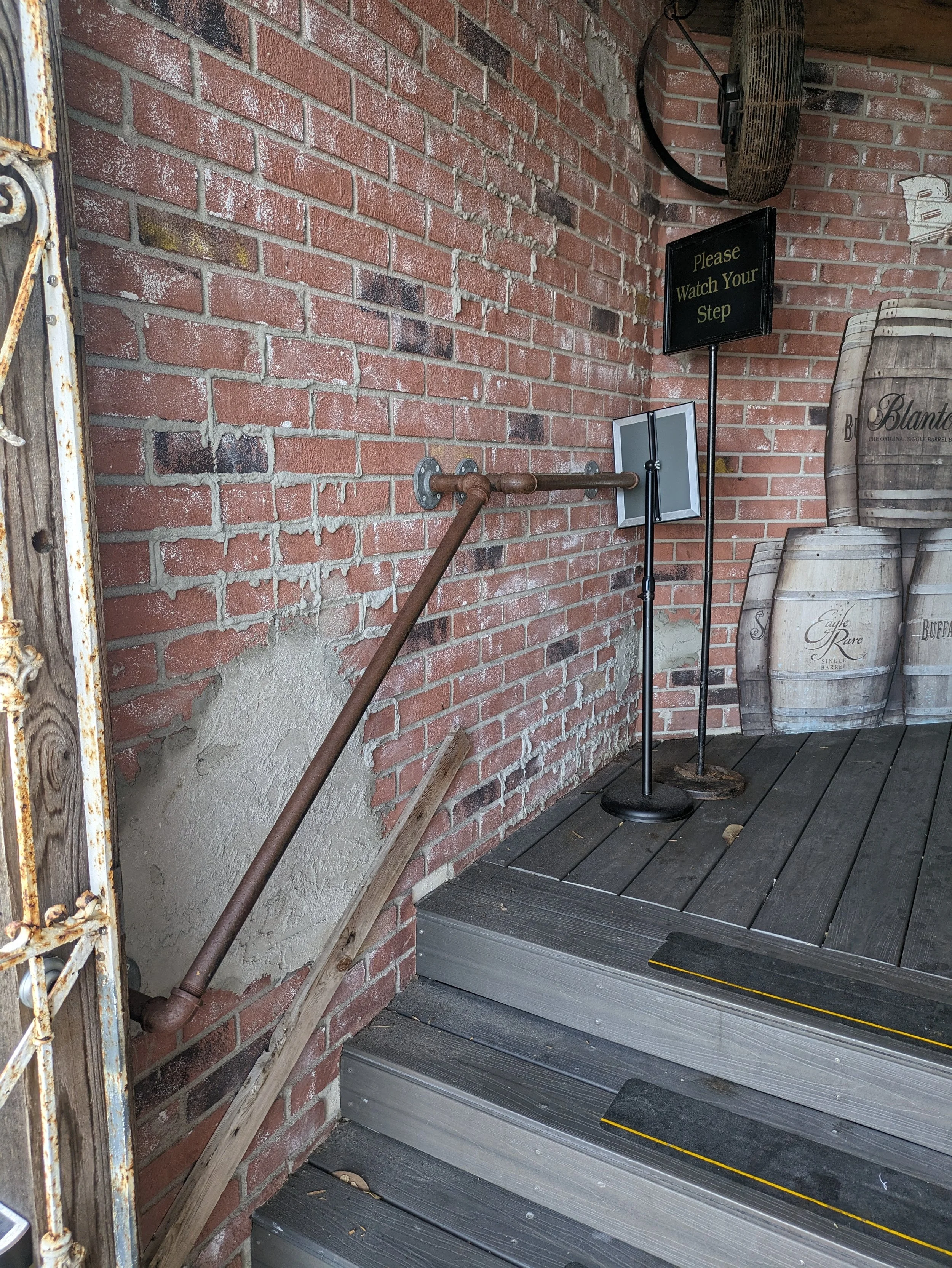 A brick wall with a rusted pipe and a wooden board leaning against the wall, next to a staircase with a yellow safety strip. A sign that says 'Please Watch Your Step' is mounted on a stand, near barrels in the background.