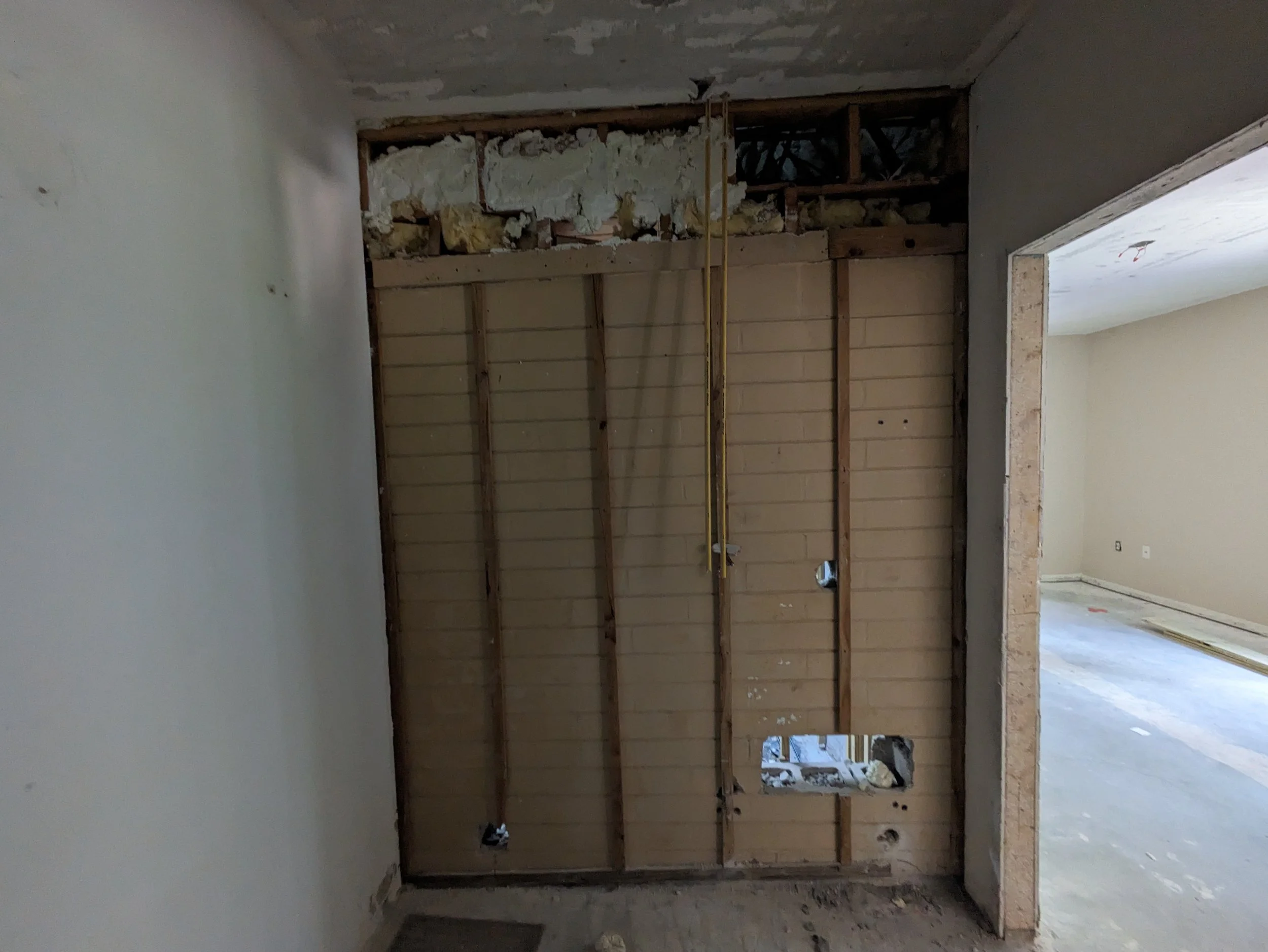 Interior view of a room under construction with a partially demolished wall, exposed framing, insulation, and electrical wiring.