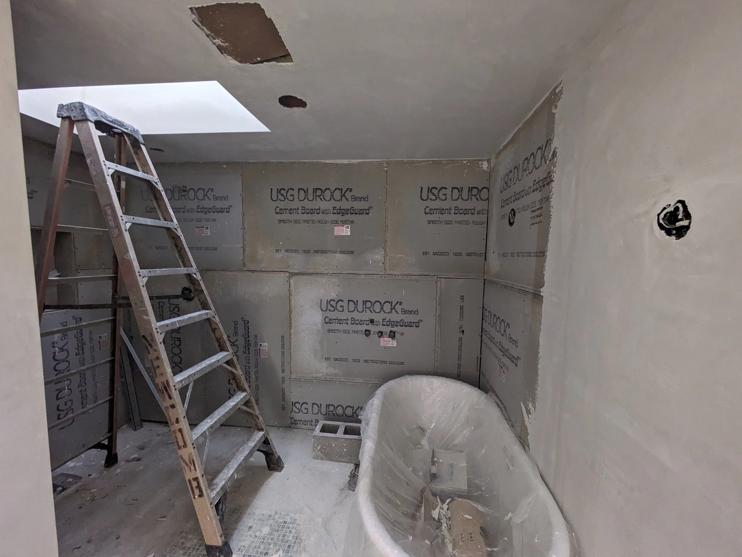 Bathroom under renovation with drywall installation, a wooden ladder, and a bathtub covered with plastic.