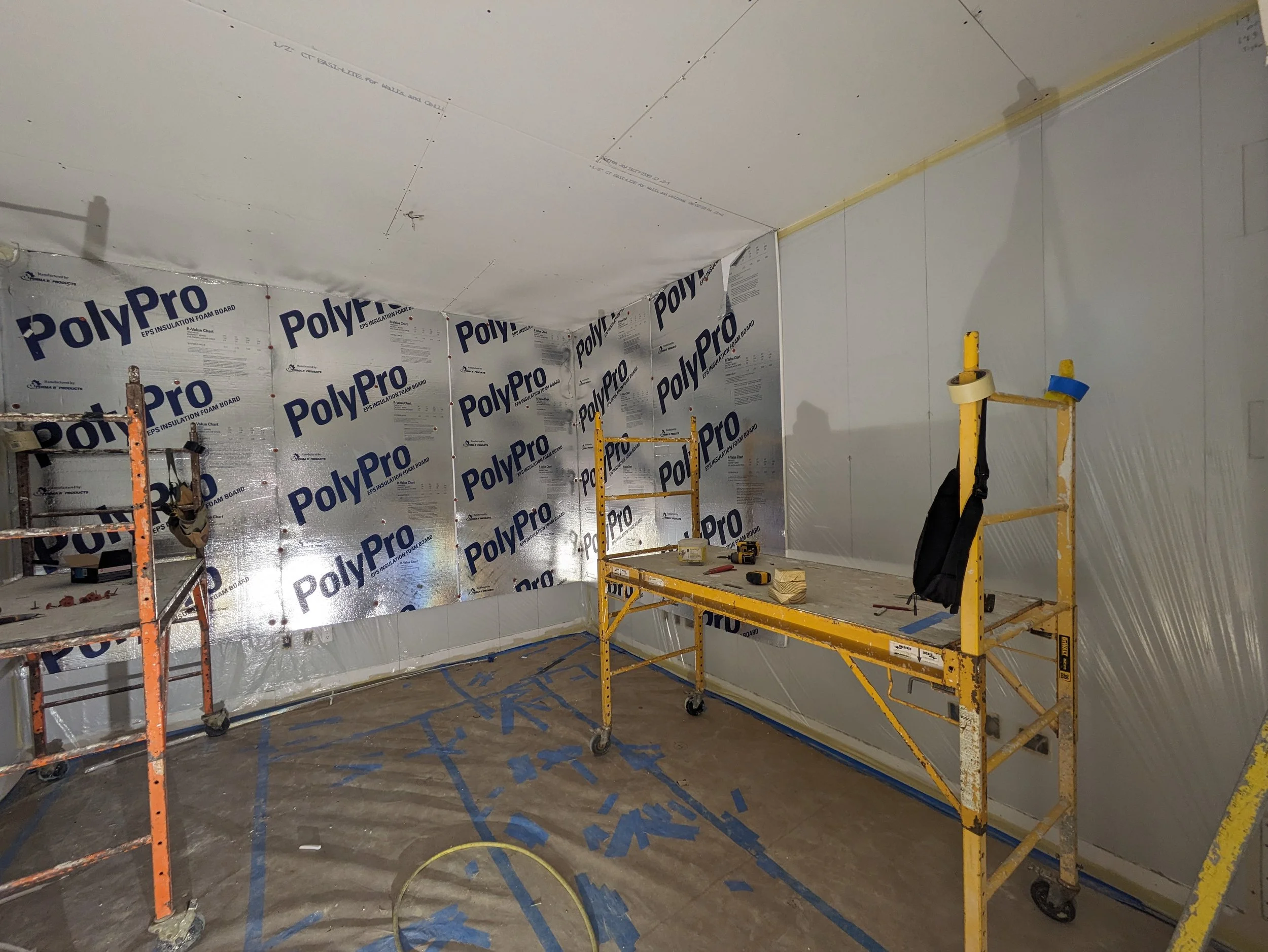 Interior of a room under construction with walls covered in PolyPro insulation boards, scaffolding, and tools on a work table.