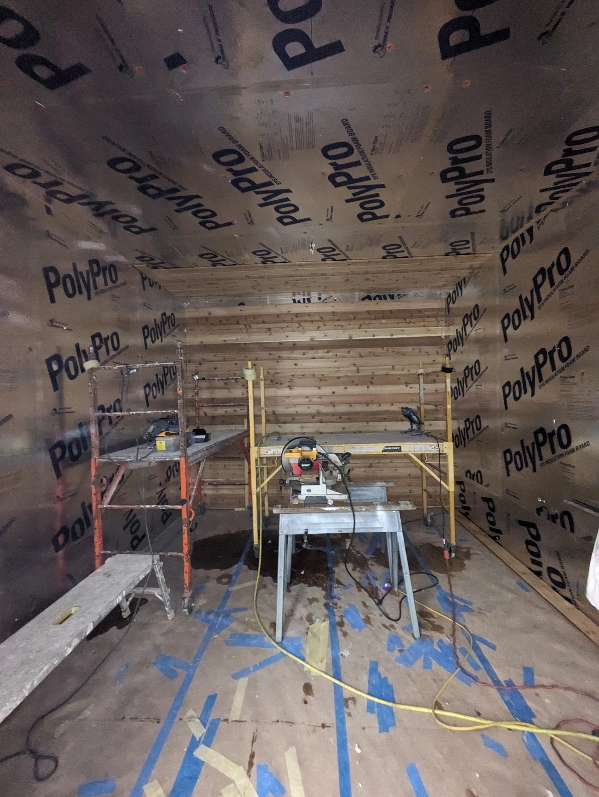 Construction room with PolyPro insulation on the walls, a work table with power tools, a ladder, and a wet floor with tape markings.