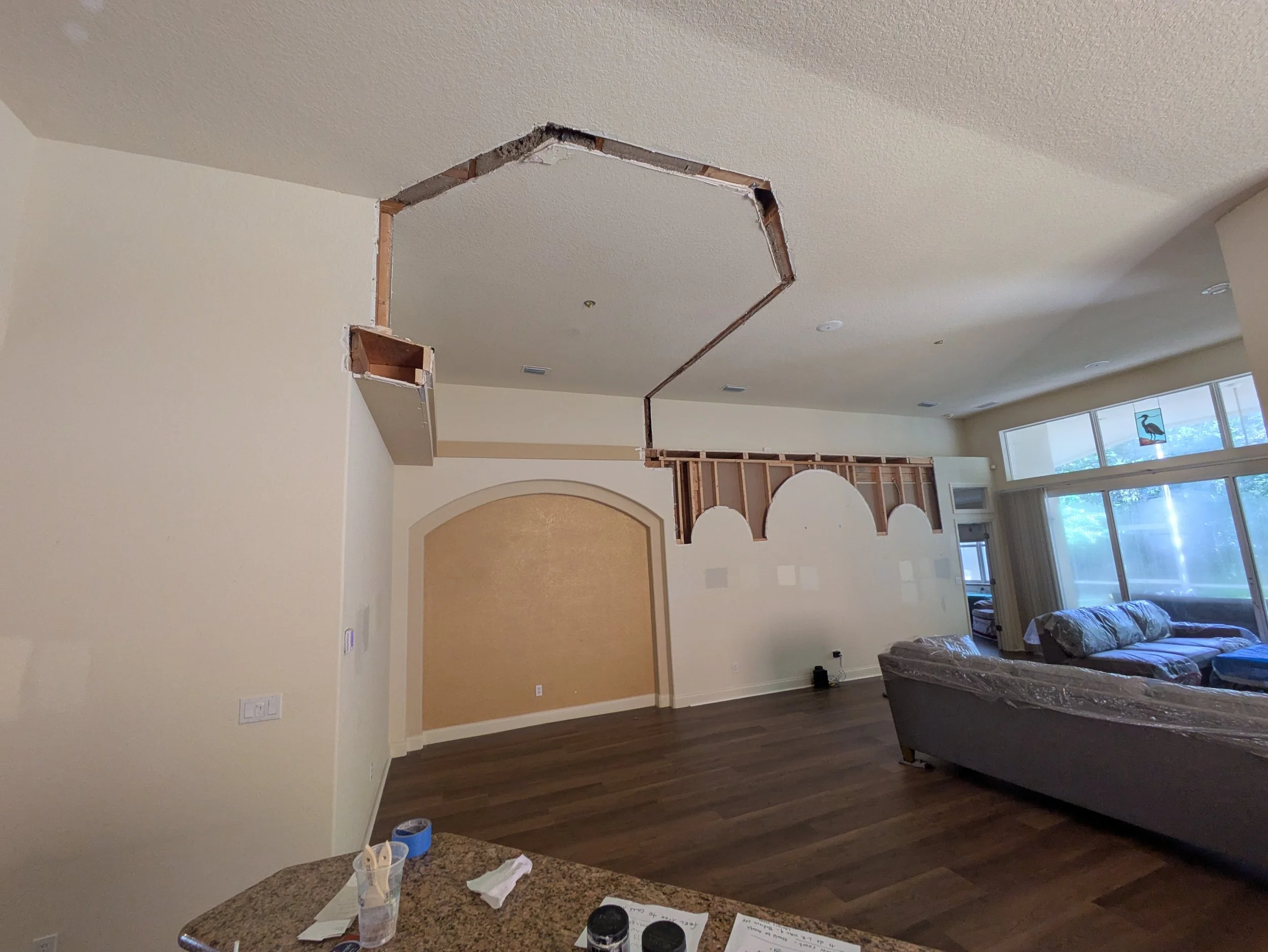 Living room with a large octagonal hole cut into the ceiling, exposing the ceiling's framing and pipes, with a partially finished wall and a sofa covered in plastic near large windows.