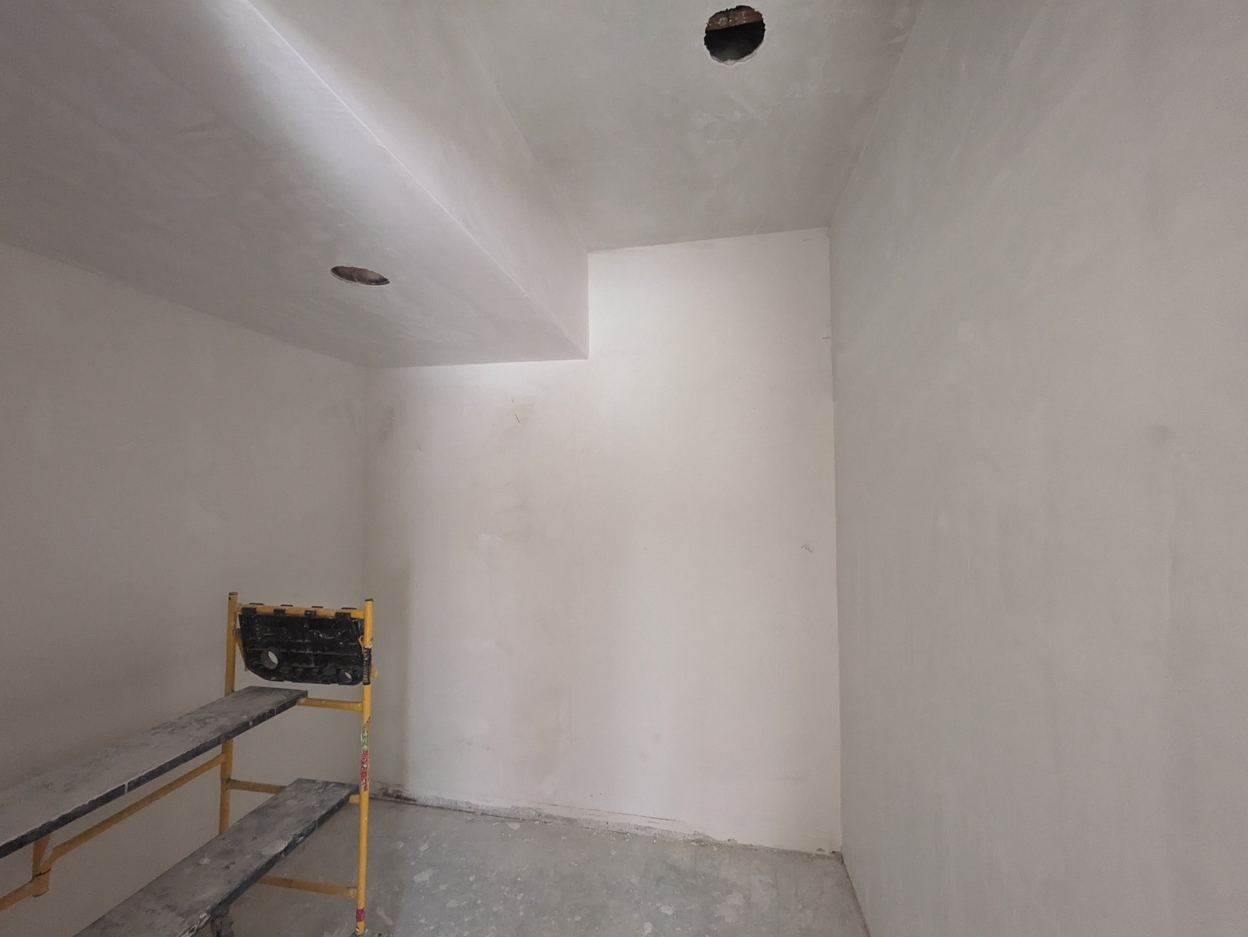 Empty room under construction with unpainted walls and ceiling, a scaffold in the corner, and overhead holes for lighting or fixtures.