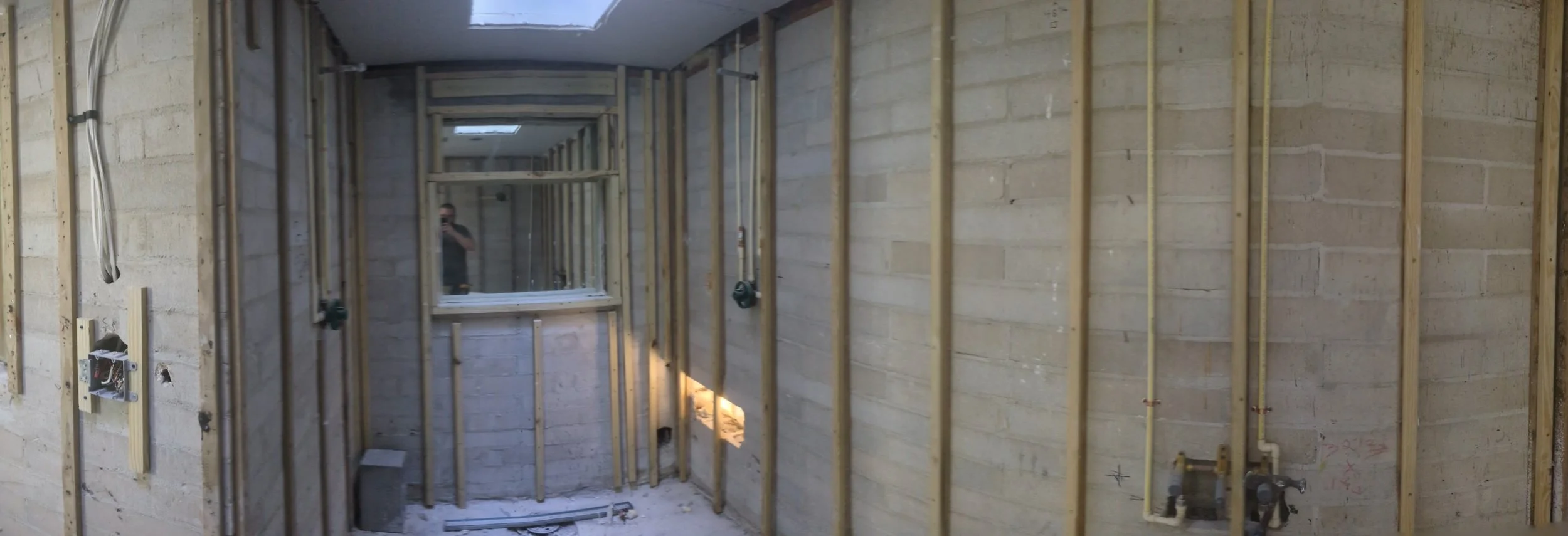 Interior of a building under construction with exposed wooden framing, electrical wiring, and plumbing, and a person taking a photo in a mirror.
