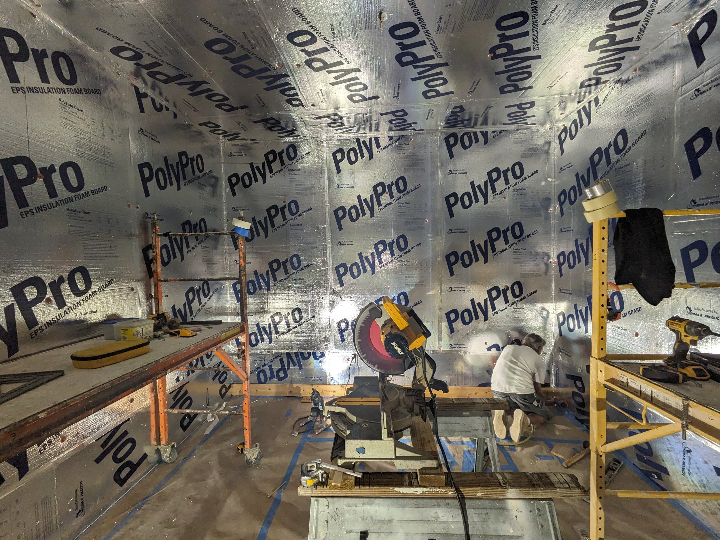 A construction worker installing insulation foam boards labeled PolyPro inside a room with silver insulation panels covering the walls and ceiling, with various tools and equipment around.