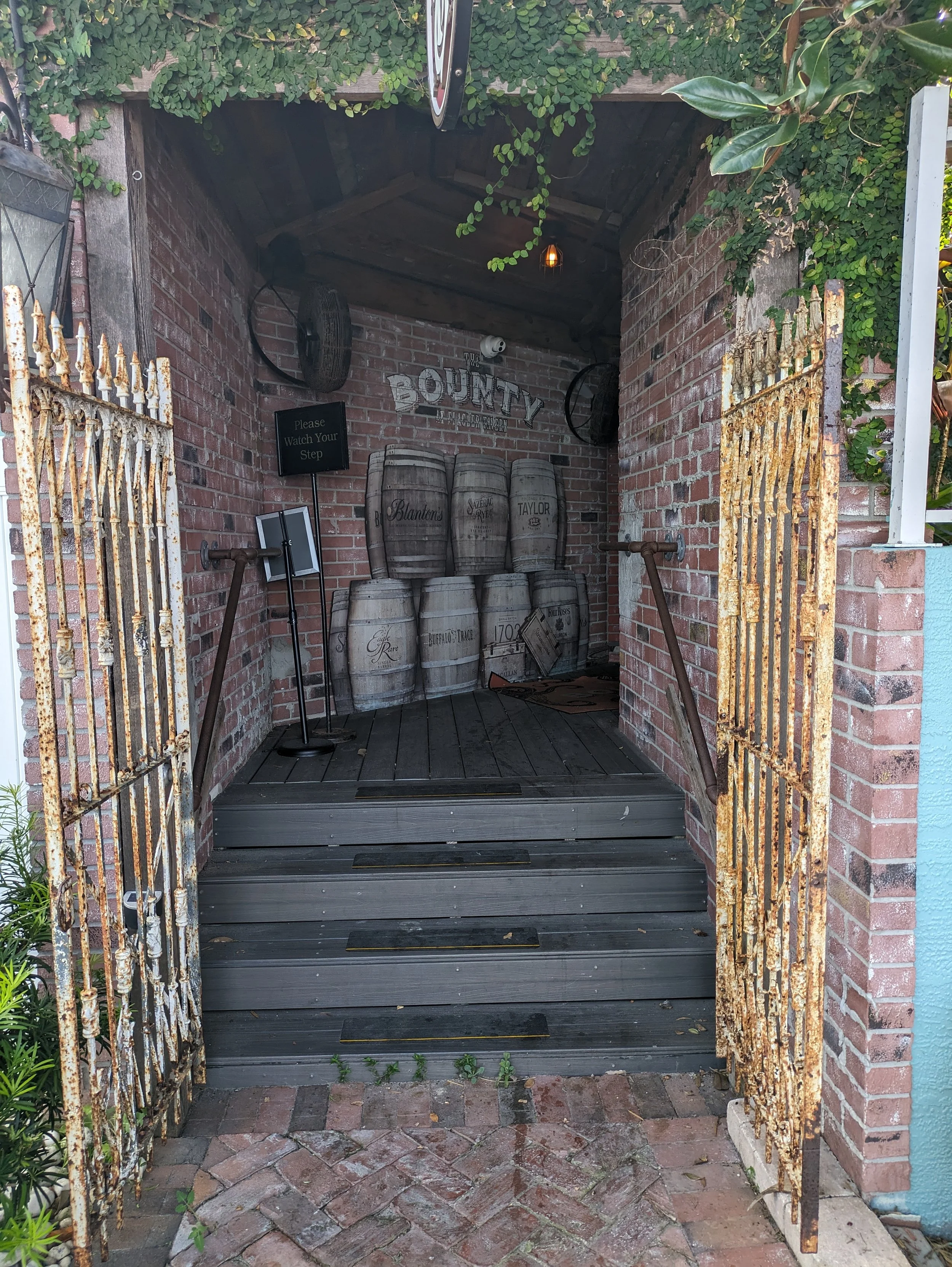 Rusty iron gates open to a small outdoor bouncypress area with stacked wooden barrels, a brick wall, a sign that says 'Please Watch Your Step', and a 'Bounty' sign on the brick wall.