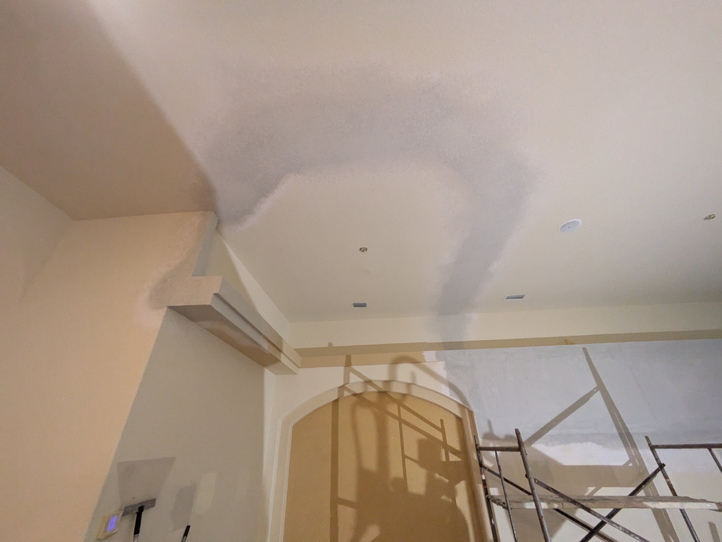Interior room with a ceiling being painted or textured, scaffolding present on the right side, beige and cream-colored walls, and a doorway with an archway.