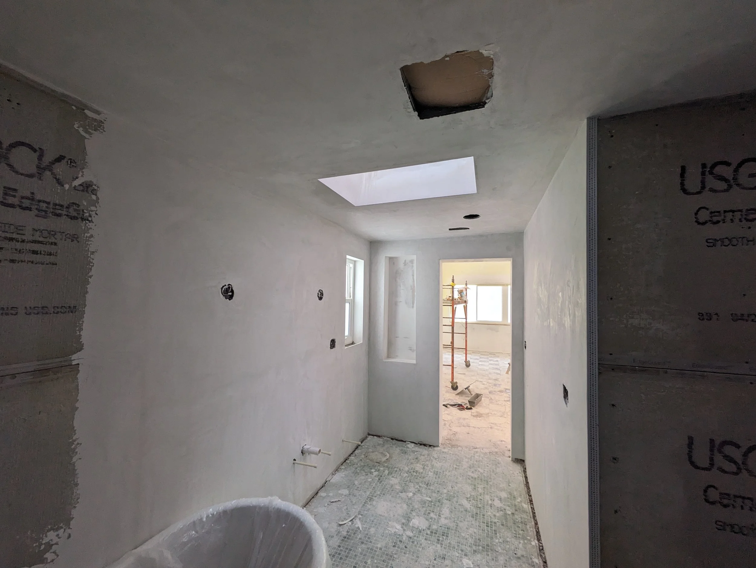 Interior of a room under construction with white walls, an opening in the ceiling, a skylight, and windows, showing ongoing drywall work.