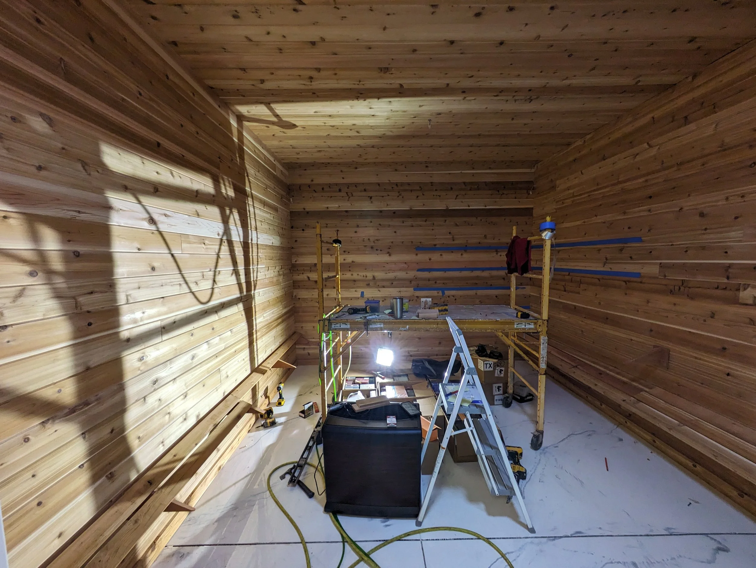 Interior of a room under construction with unfinished wooden walls, a scaffold, a ladder, and various tools and materials.