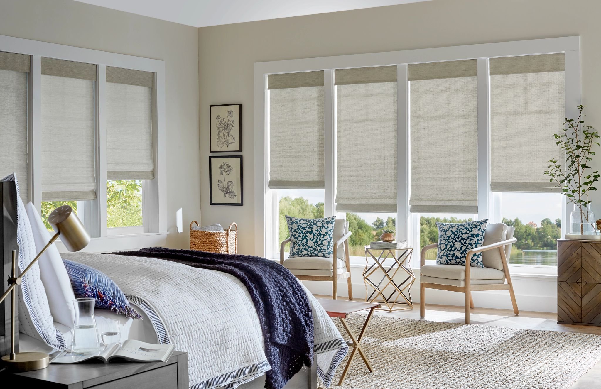 Bright bedroom with large windows covered by beige blinds, a bed with white quilt and navy throw, two chairs with blue and white cushions, a side table with a stack of books, a chair, and houseplants near a lake view