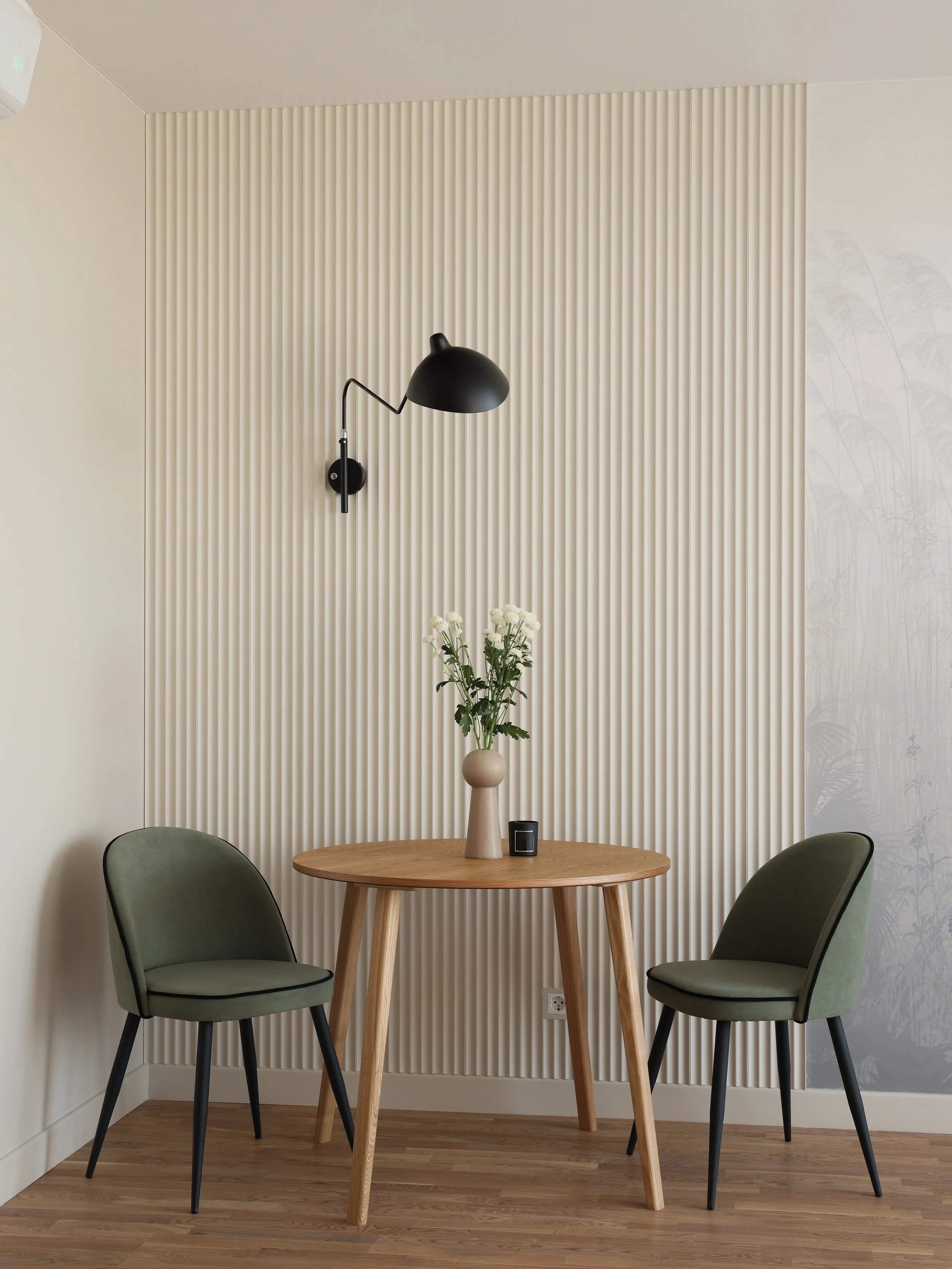 A small round wooden table with a bouquet of white flowers in a beige vase and a small black object. Two green upholstered chairs with black legs are placed on either side of the table. A black wall-mounted lamp is on a striped beige and white textured wall, with a light wood floor.
