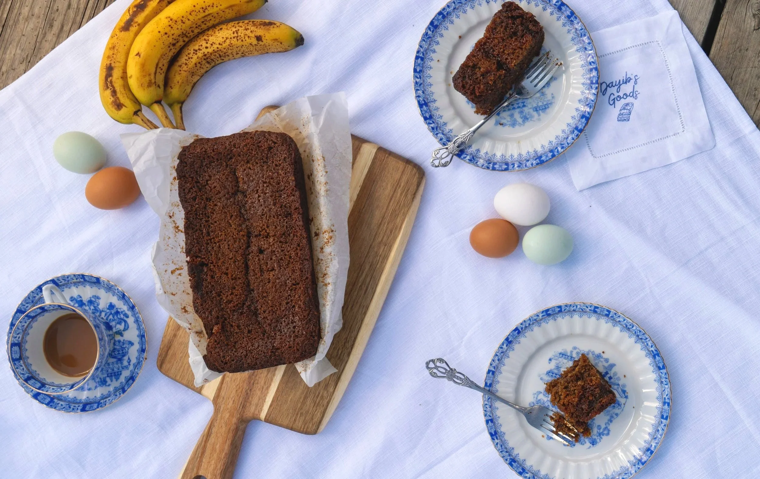 Bananas, eggs, a loaf of banana bread, a slice of banana bread on a blue and white plate, a cup of coffee, and a napkin with embroidered text on a white tablecloth.