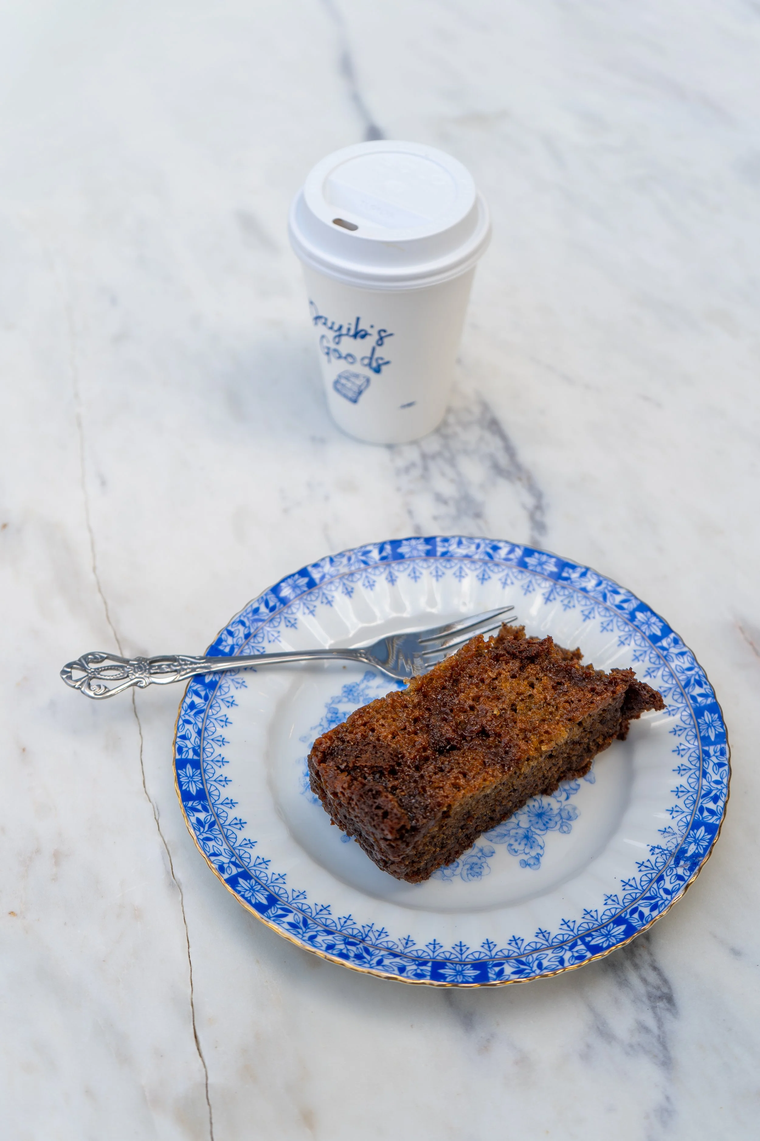 Slice of butterscotch banana bread on a decorative blue and white plate with a silver fork, accompanied by a white to-go coffee cup on a white marble surface.