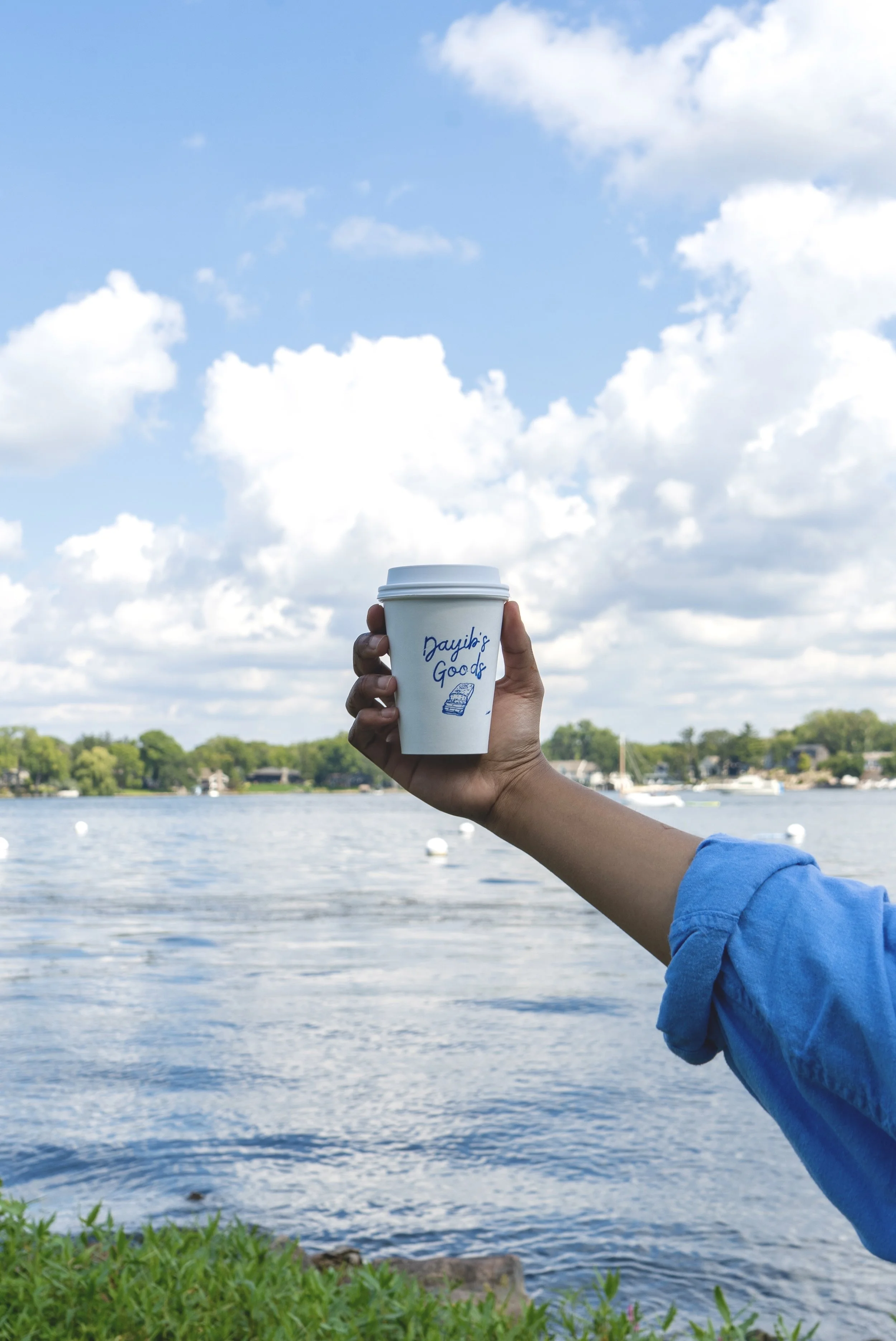 Hand holding a disposable Dayib's Goods coffee cup with a lake, trees, boats, and a blue sky with white clouds in the background.